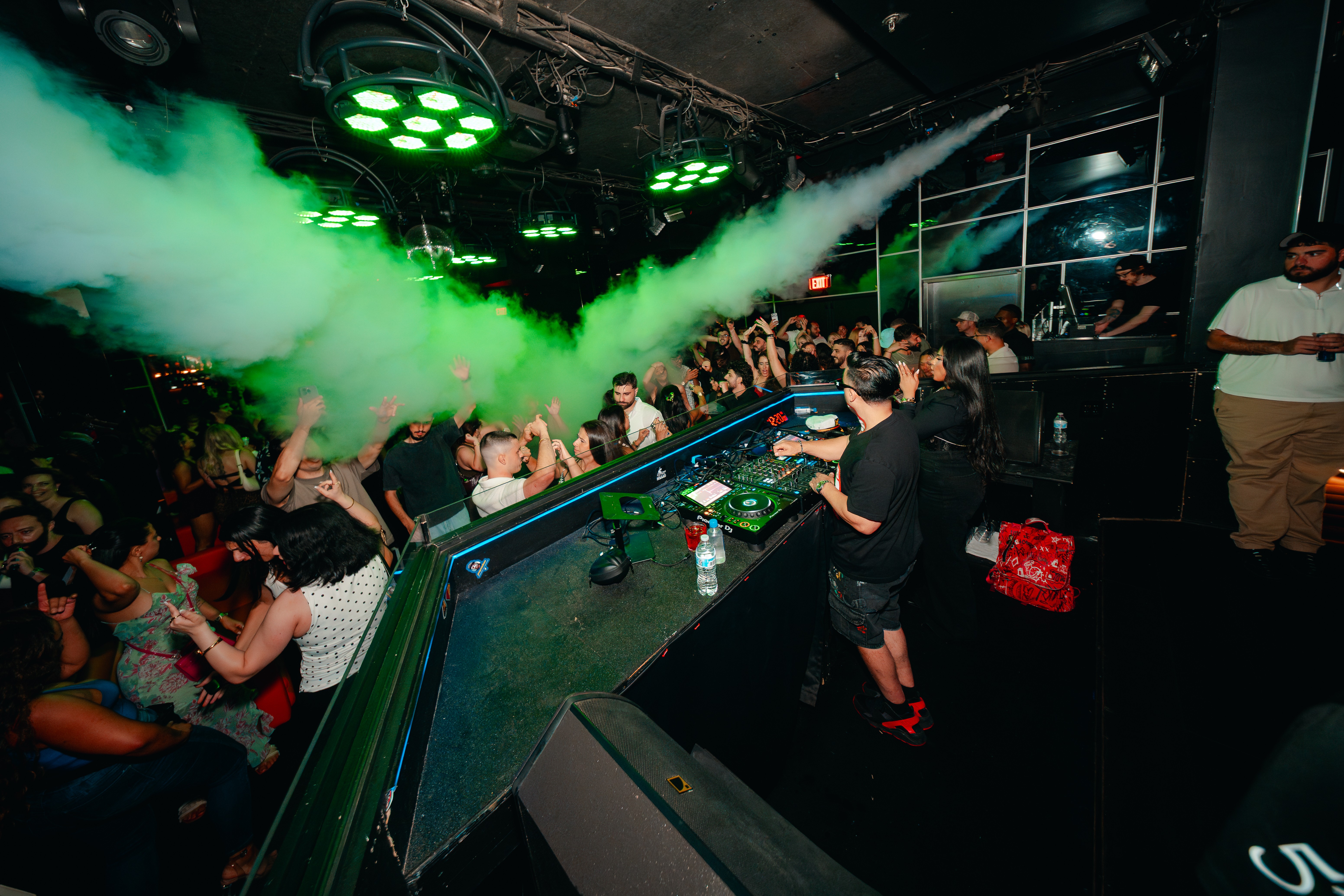 Nightclub scene with green laser lights, smoke, and a crowd dancing and socializing, viewed from the DJ booth area.