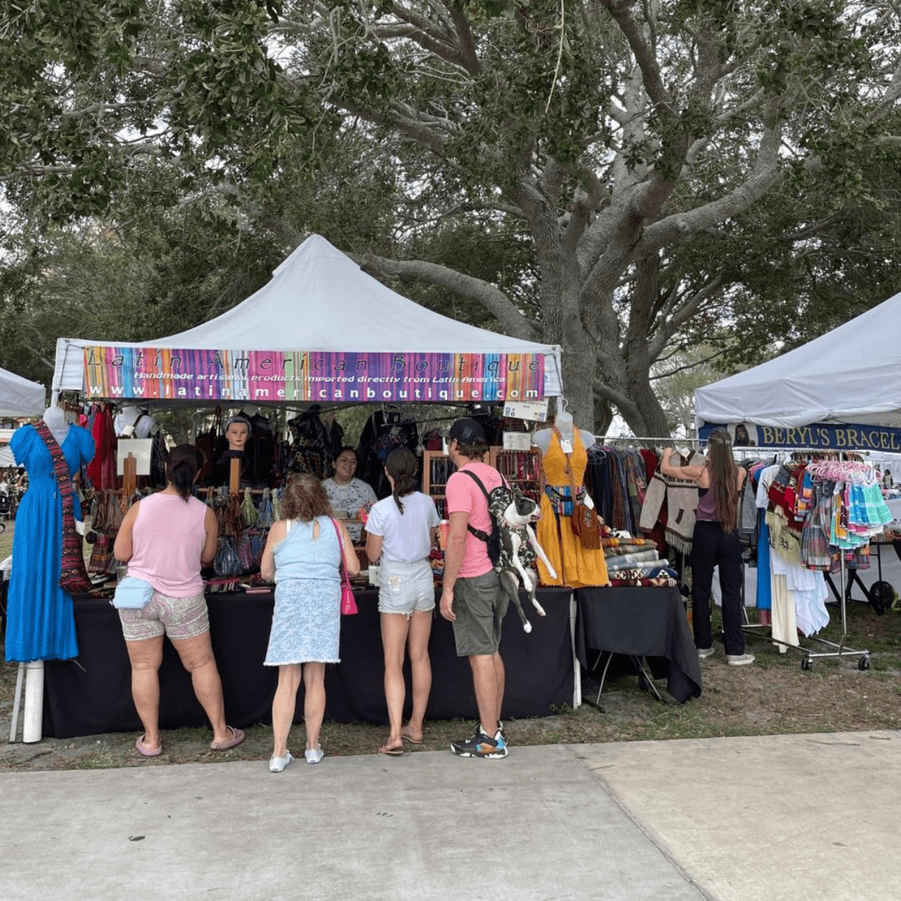 People browsing outdoor market stalls with colorful fabrics and clothing, trees in the background, under tents.