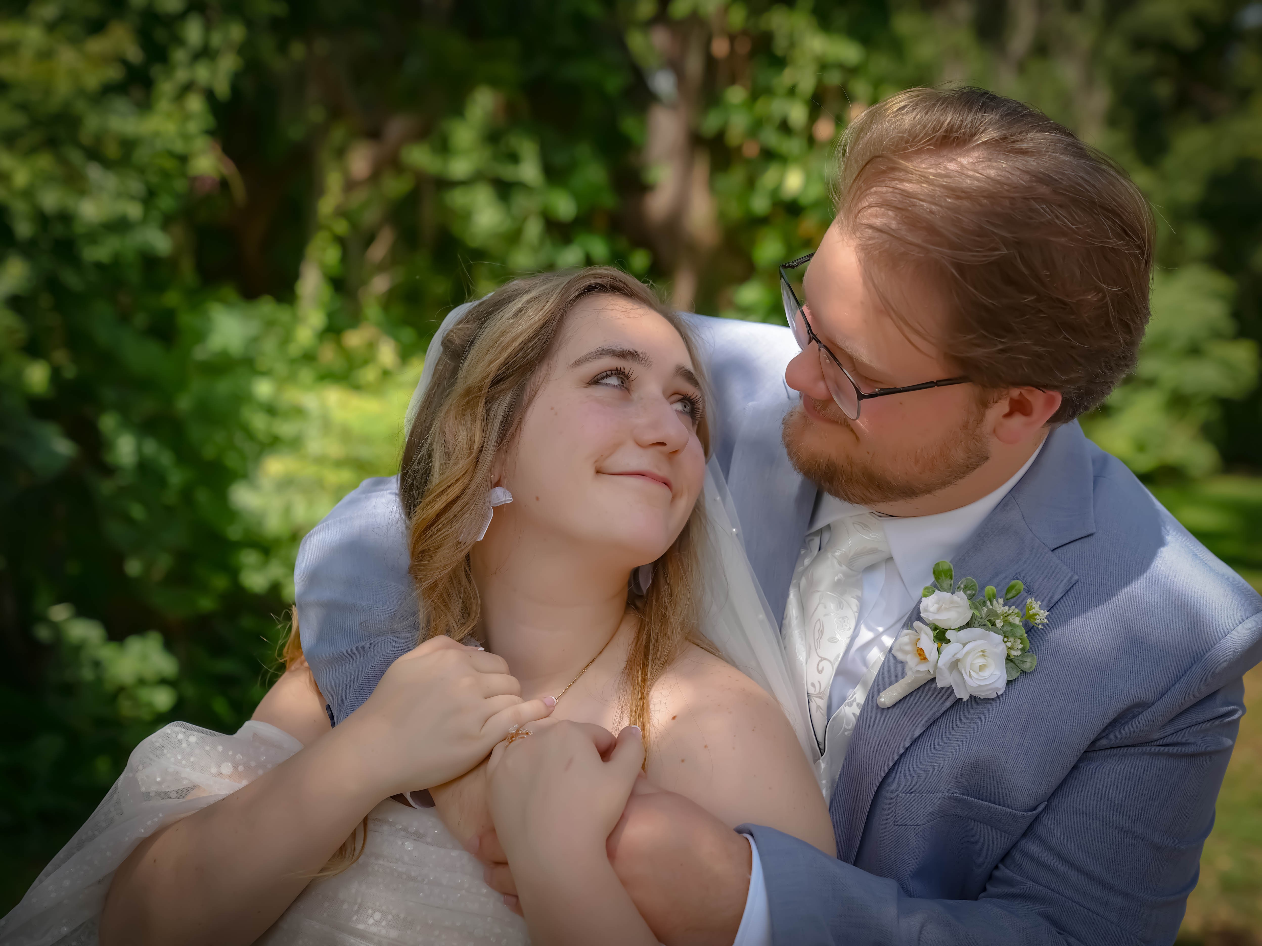 A bride and groom outdoors, smiling and looking at each other, surrounded by green trees.