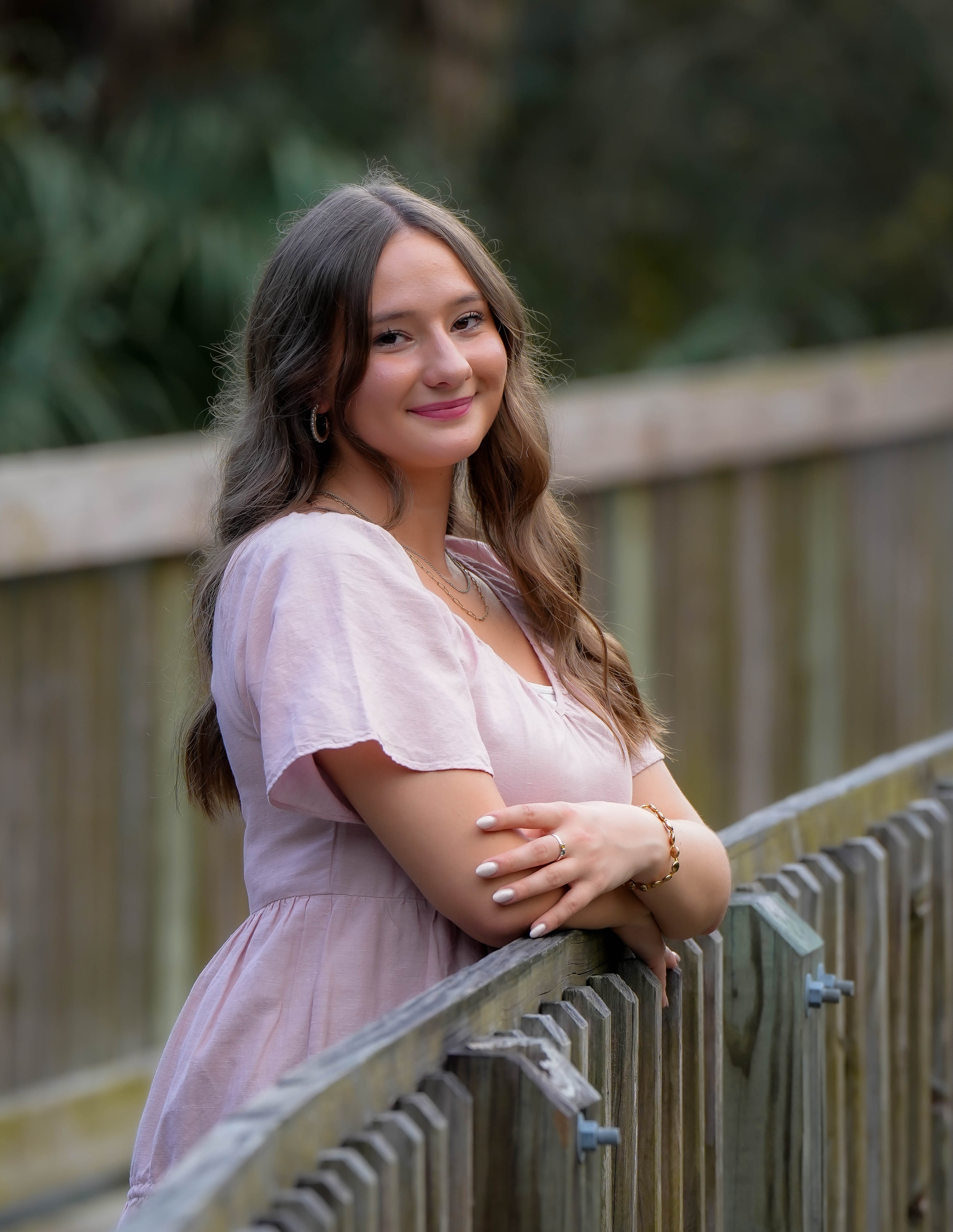 Young woman with long wavy hair smiling outdoors, leaning on a wooden fence, wearing a light pink dress.