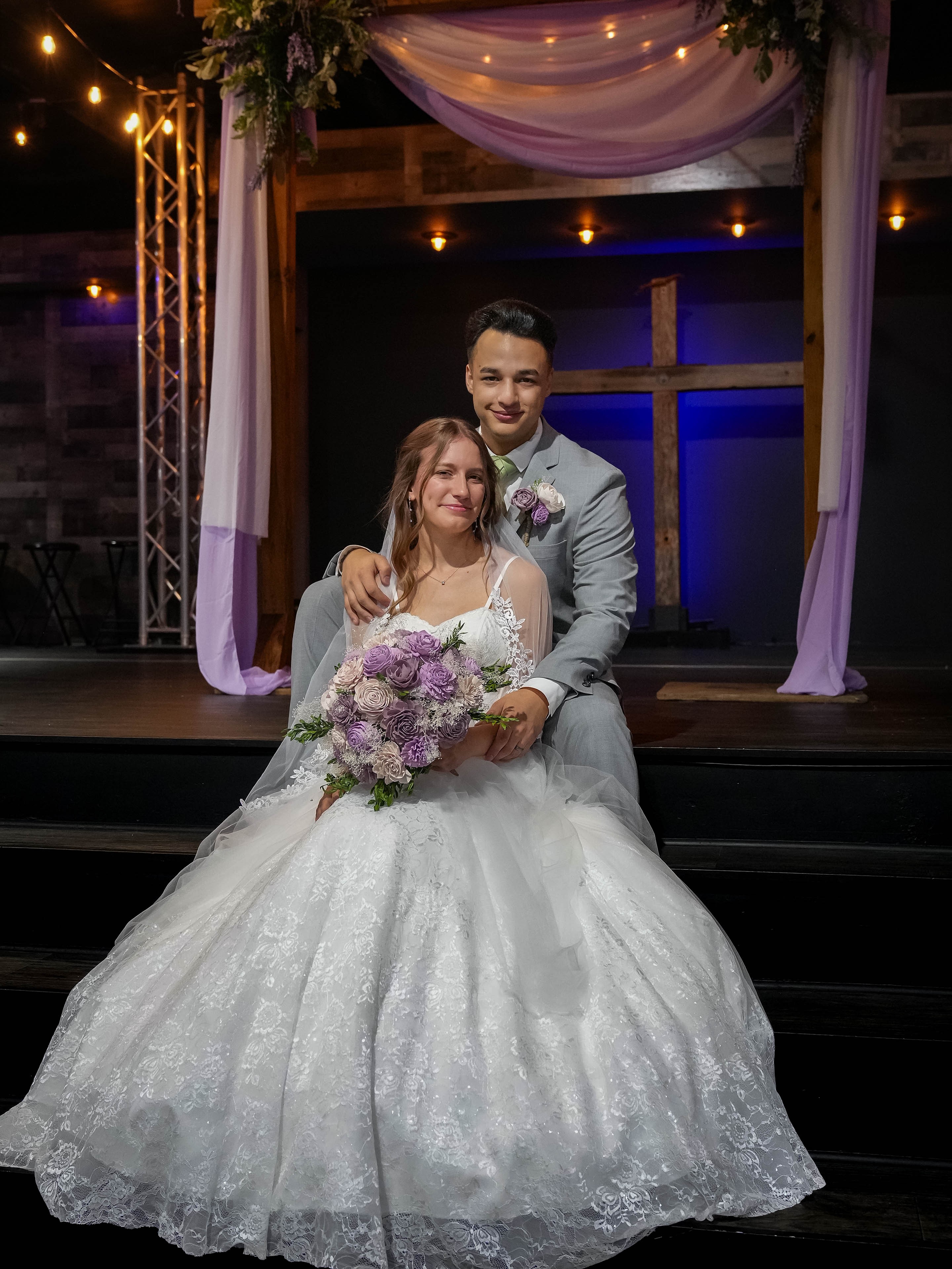 Bride in a white wedding gown holding a bouquet, seated on stairs with groom standing behind her, stage with drapes and lights in background