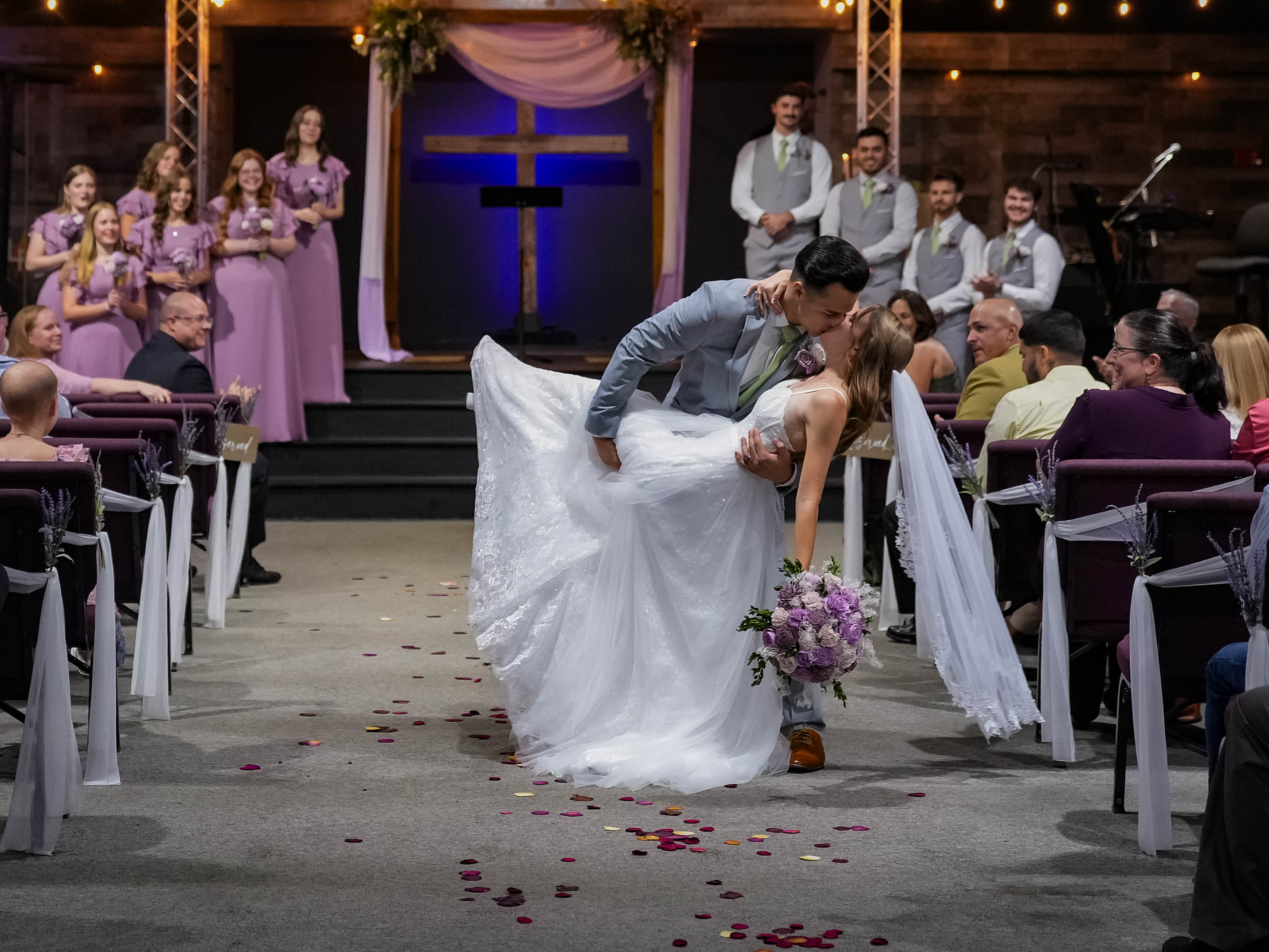 Bride and groom kiss in the aisle during wedding ceremony, guests seated on both sides, choir in purple dresses in background.