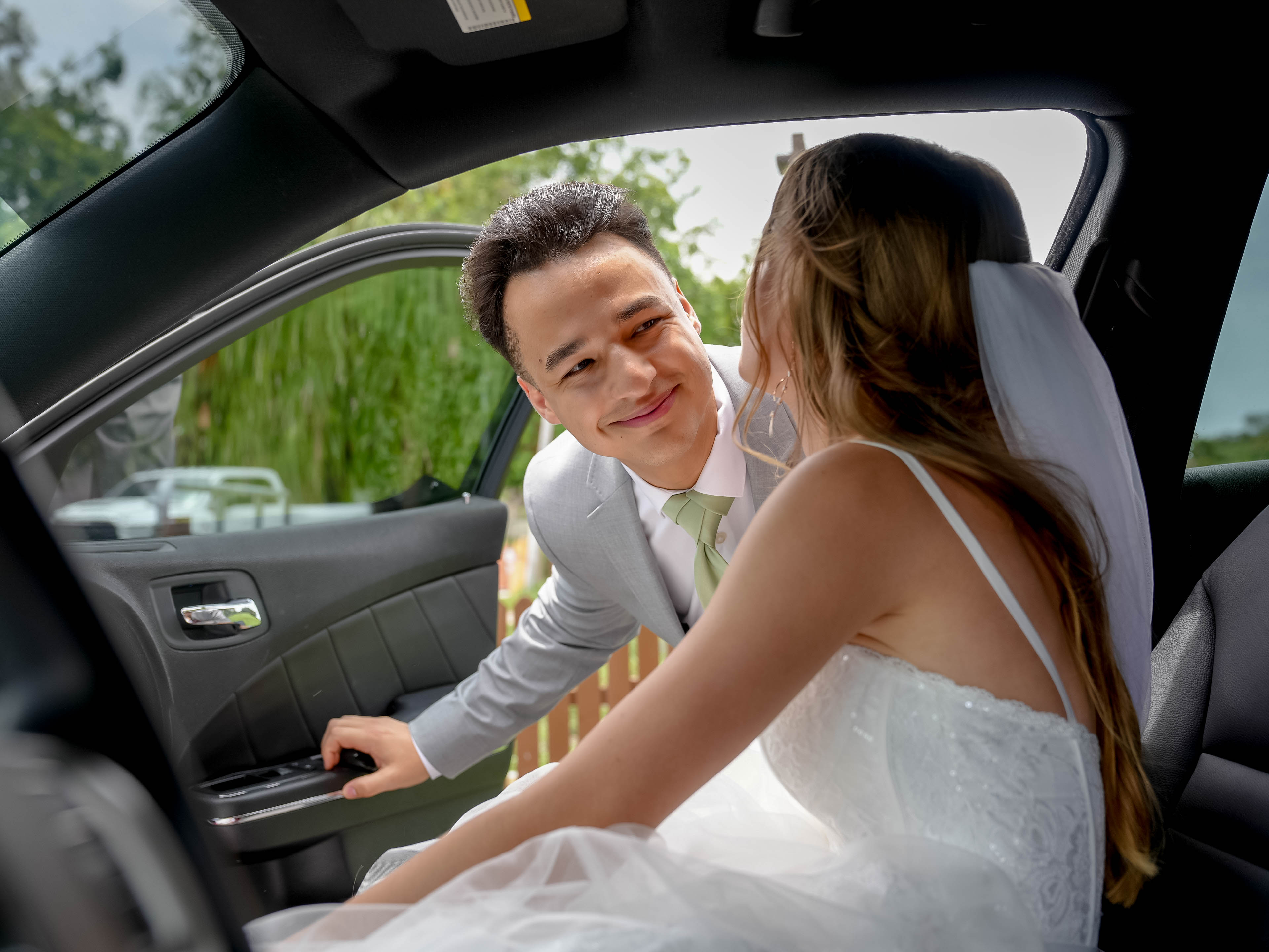 A bride and groom inside a car, smiling at each other, with the groom leaning in towards the bride.