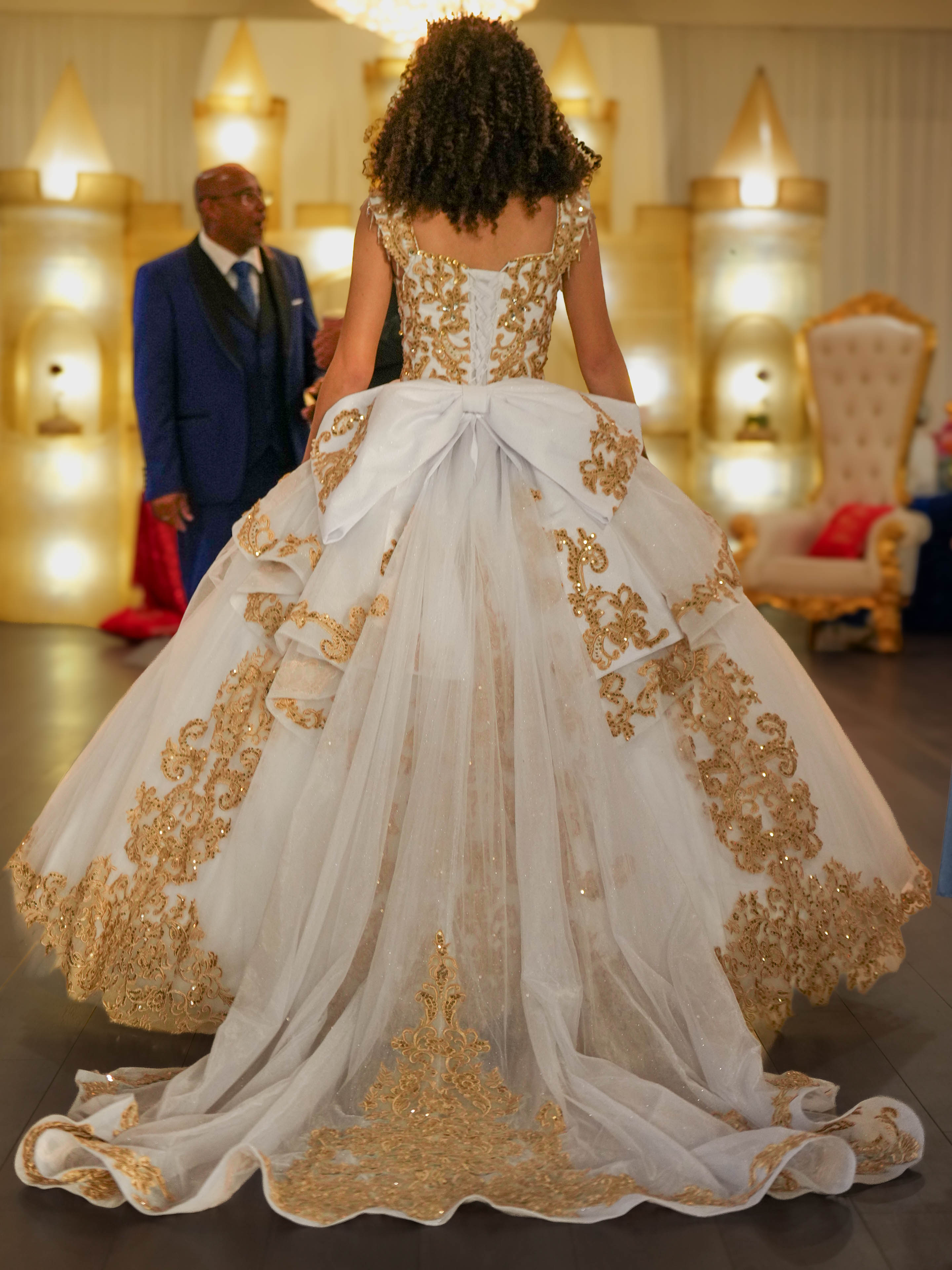 Woman in an ornate wedding gown with gold embroidery, standing in a decorated room with two men in the background.