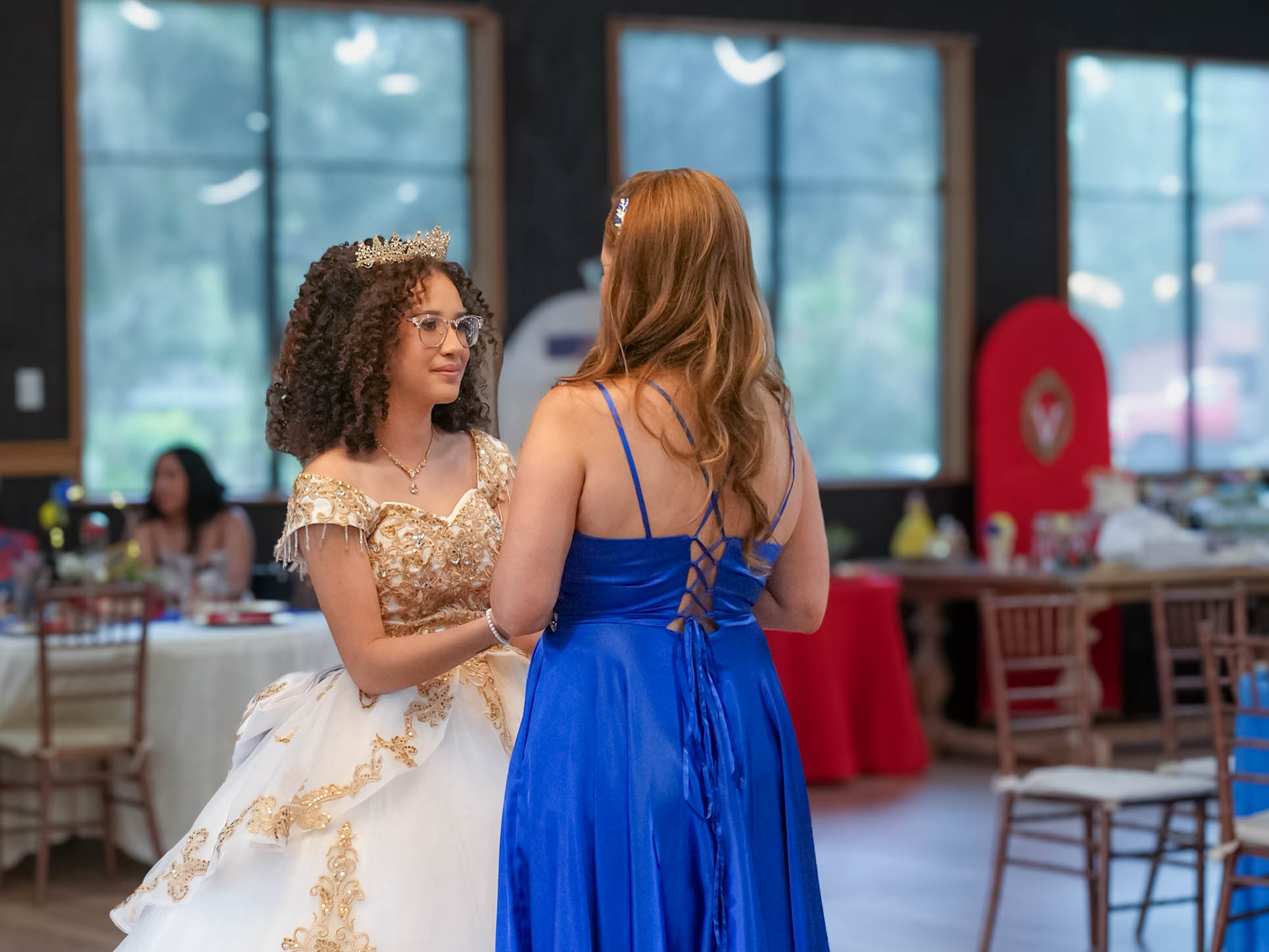 Two women in formal dresses talking indoors, with tables and large windows in the background.