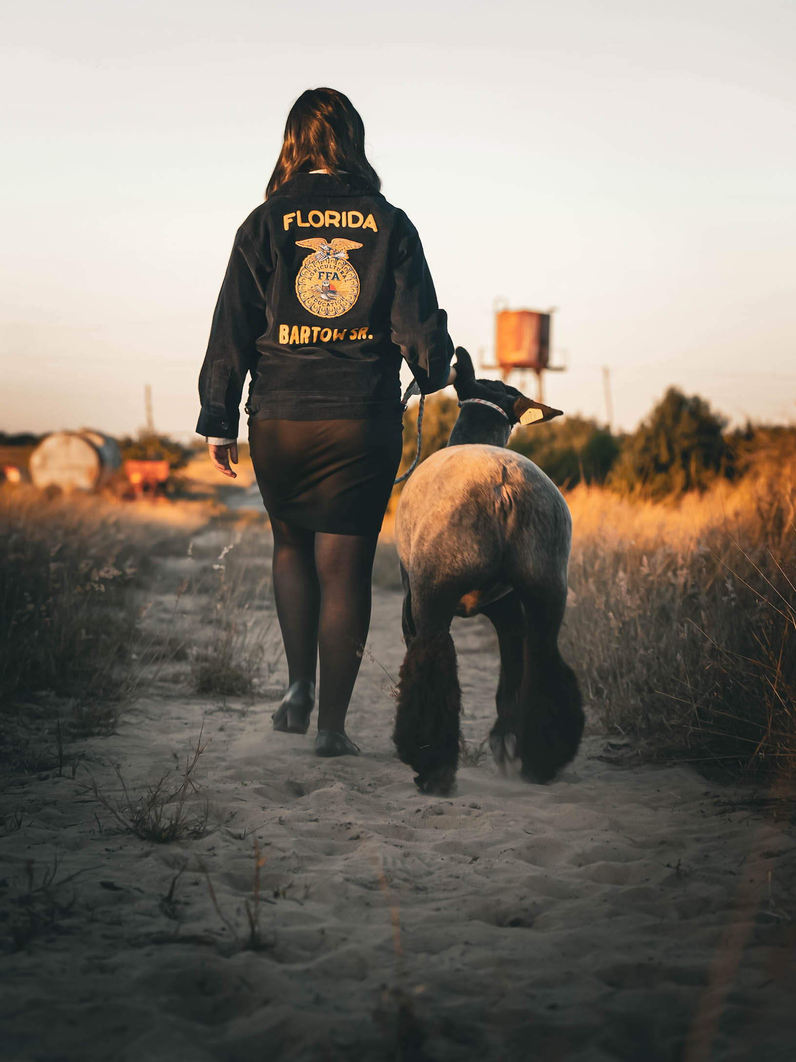 Person walking on a dirt path with a dog, wearing a black jacket with Florida and Bartow, Florida text, and a skirt.