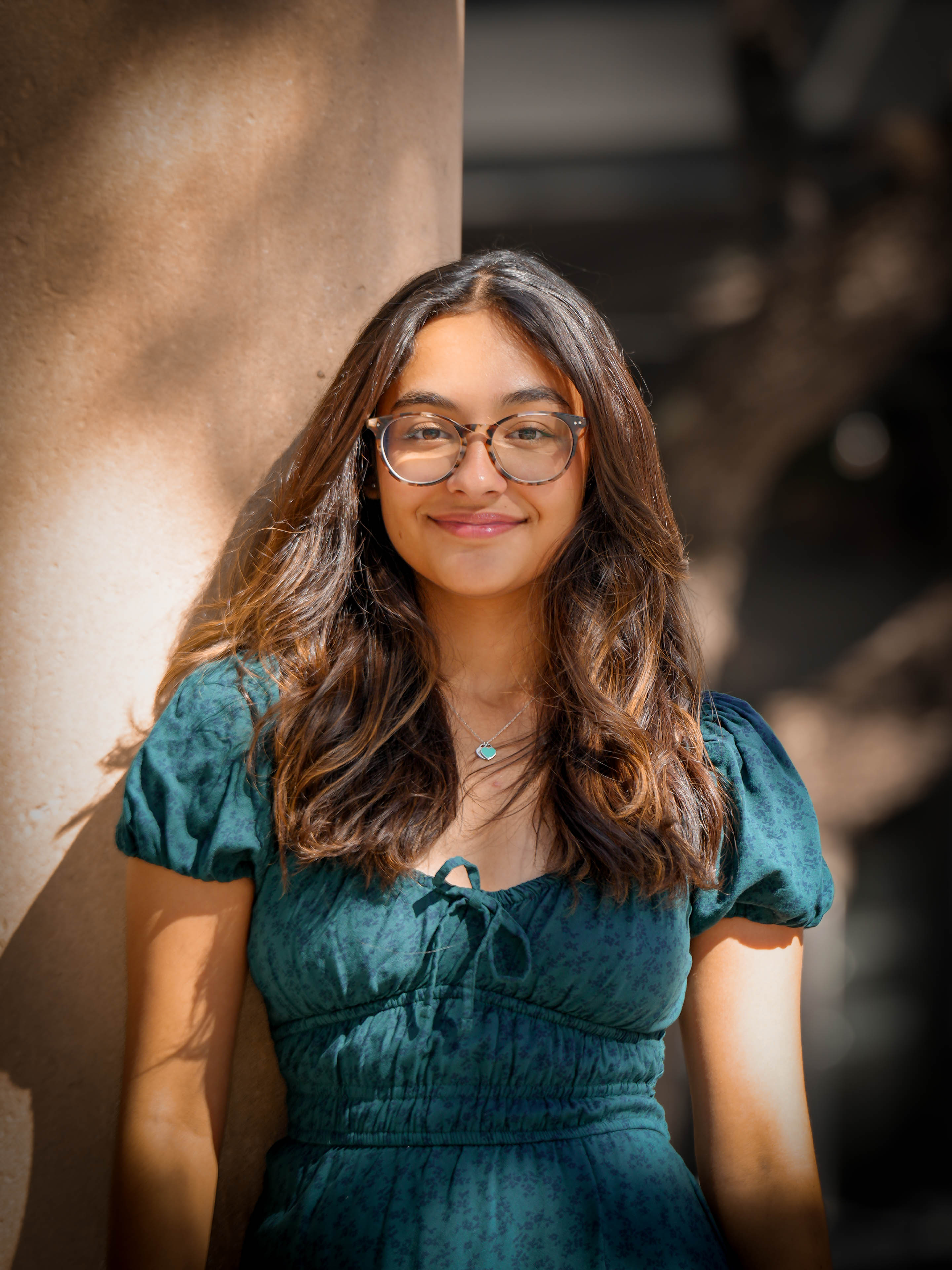 Young woman with glasses and long wavy hair smiling outdoors, wearing a teal dress, standing near a wall.