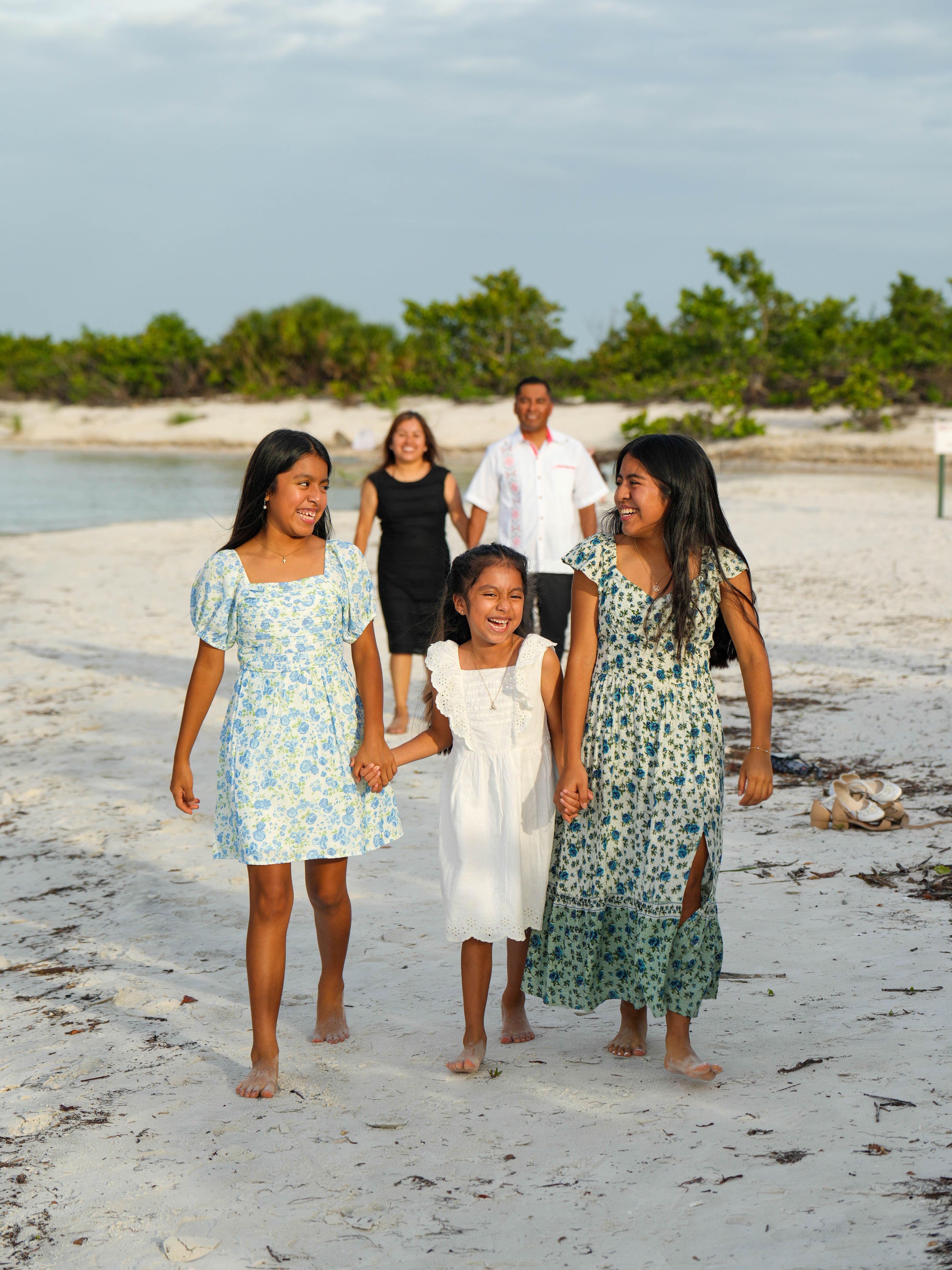 Group of five people walking on beach, two adults and three children, with trees and sky in background.