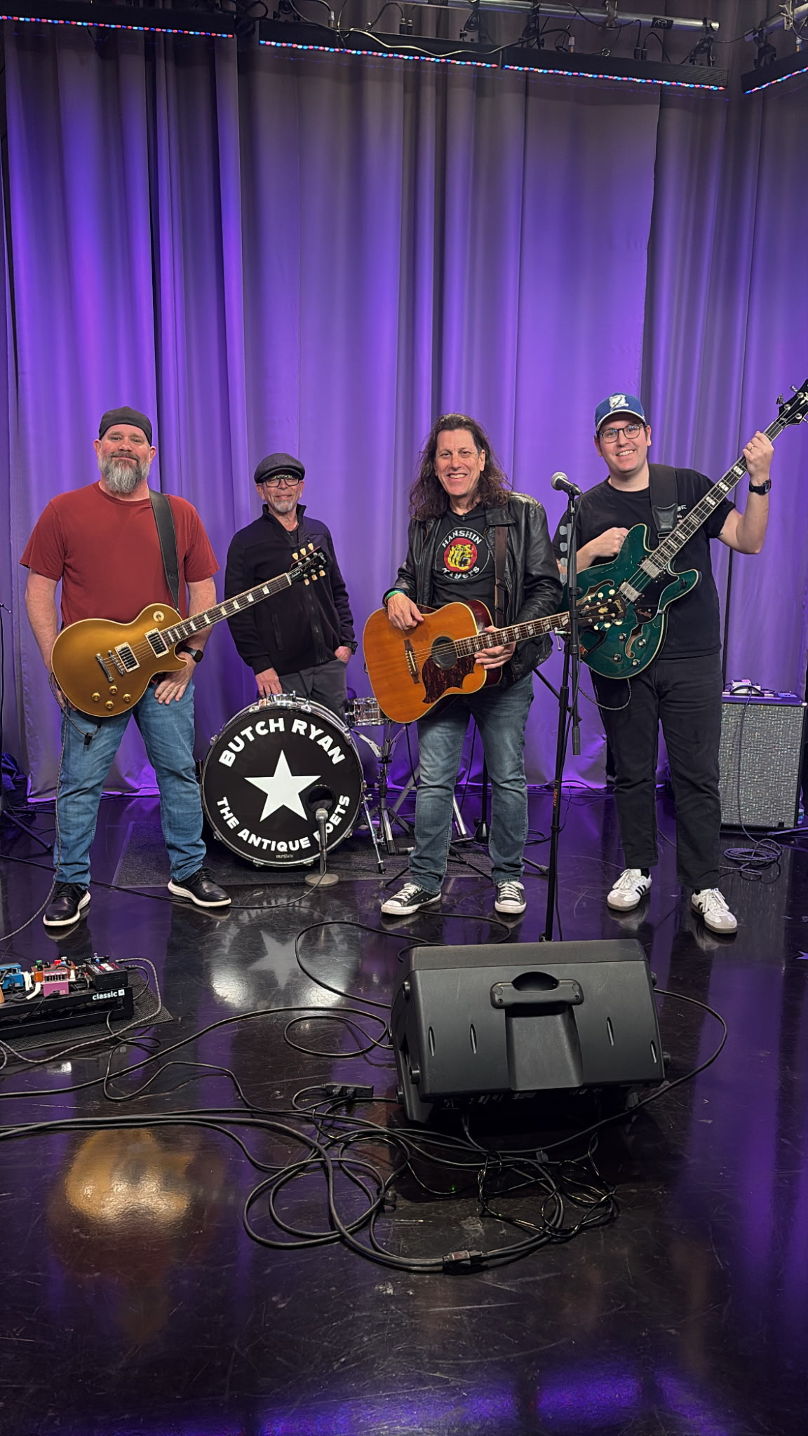 Four musicians on stage with purple curtains, holding guitars and a drum, smiling at the camera.