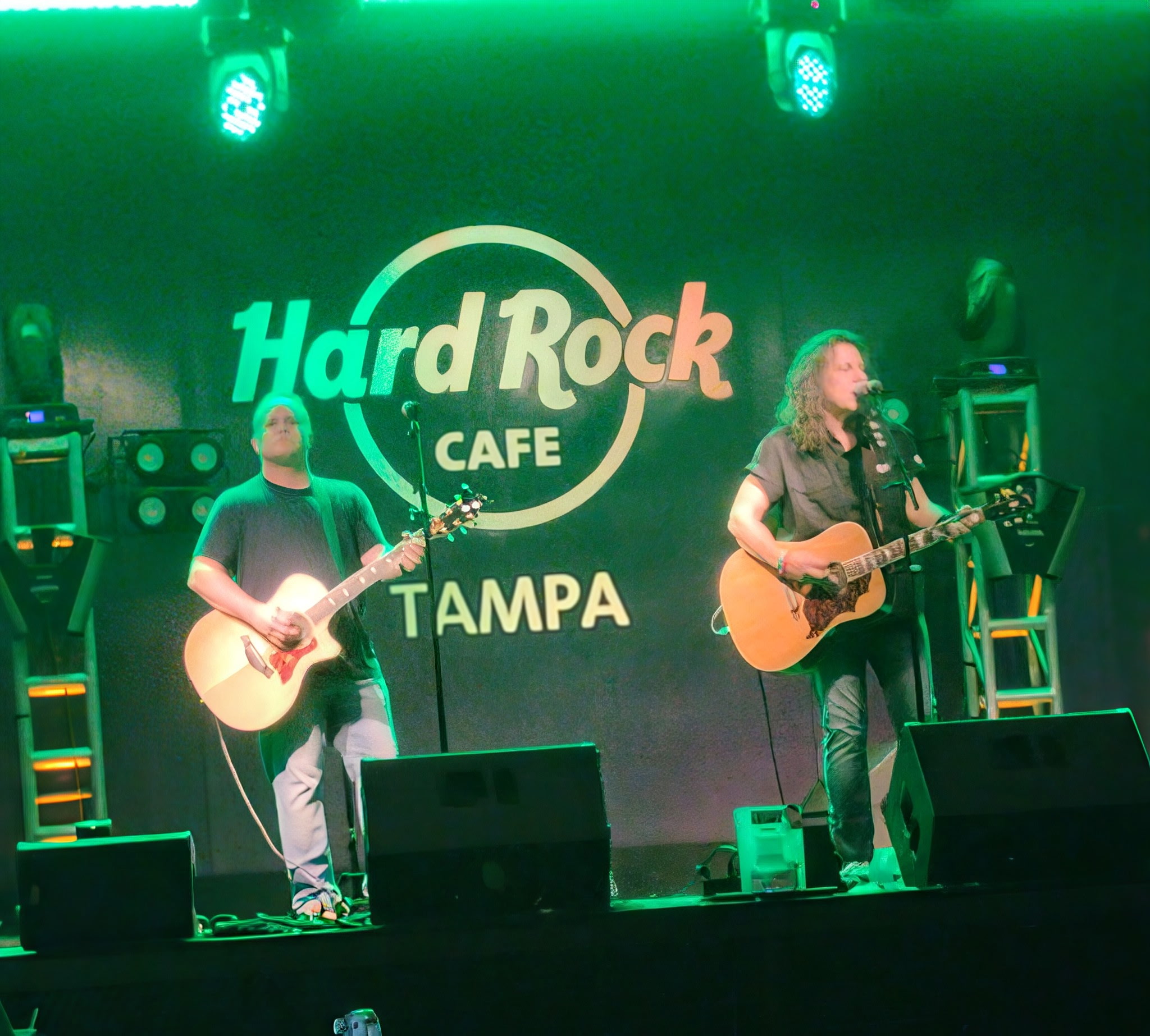 Two musicians perform on stage with guitars, a large Hard Rock Cafe Tampa logo in the background, and stage lighting.