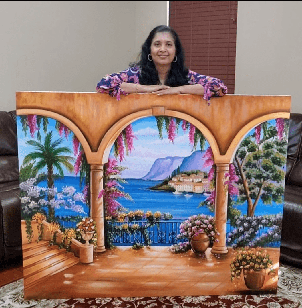 Woman smiling behind a colorful painted backdrop of a seaside landscape with trees, flowers, and a small village.