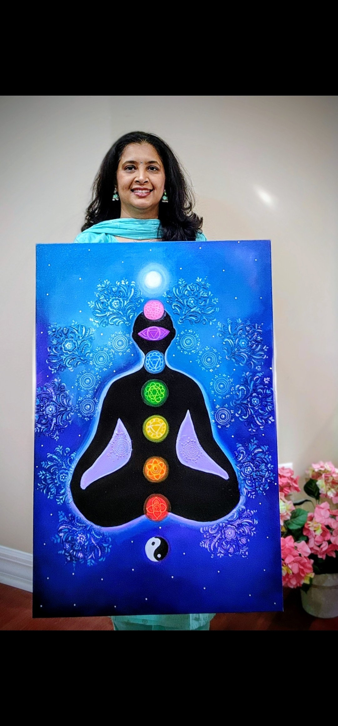 Woman holding a colorful painting of a person in a meditative pose with chakras, against a blue background, smiling.