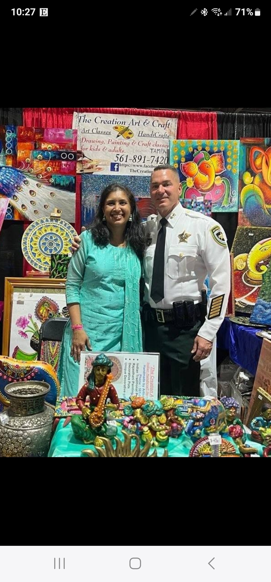 Two people, a woman and a man in uniform, standing in front of colorful artwork and crafts at an event.