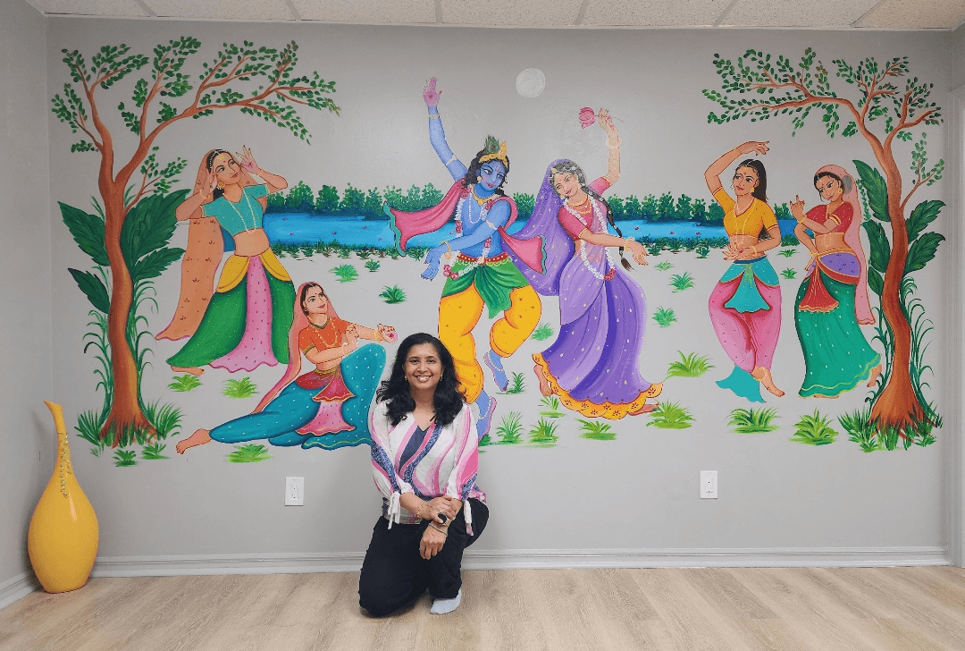 Woman kneeling in front of a colorful mural depicting mythological figures in a landscape with trees and water.