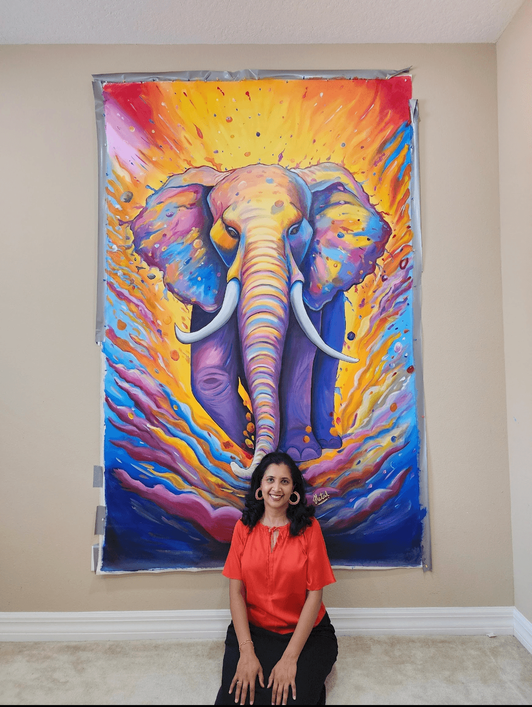 Woman sitting in front of a colorful elephant painting on a wall, with vibrant yellow, orange, blue, and purple hues.