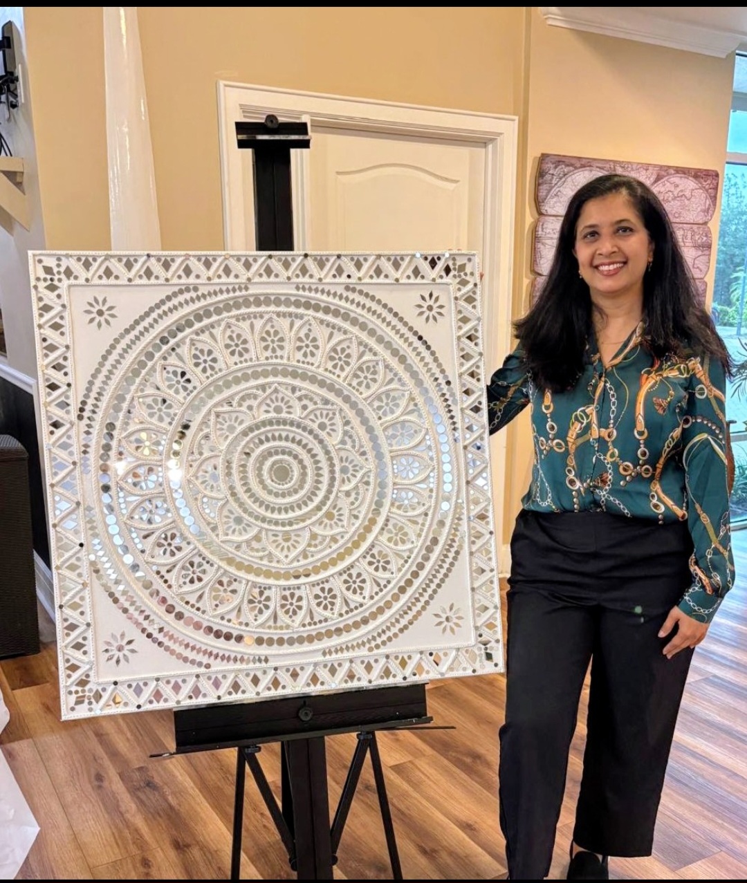 Woman standing next to a large, intricate circular artwork on an easel, smiling indoors with wooden flooring and beige walls.