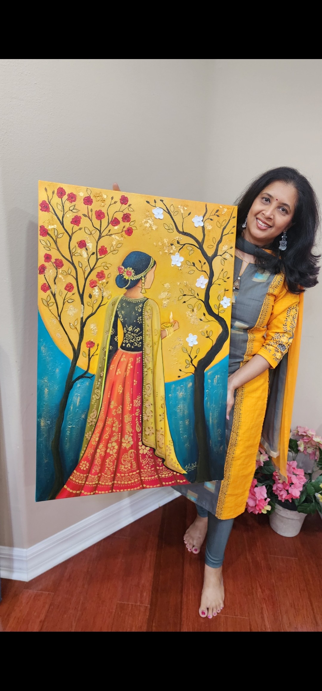 Woman standing next to colorful painting of a woman in traditional attire, with flowers nearby, indoors.
