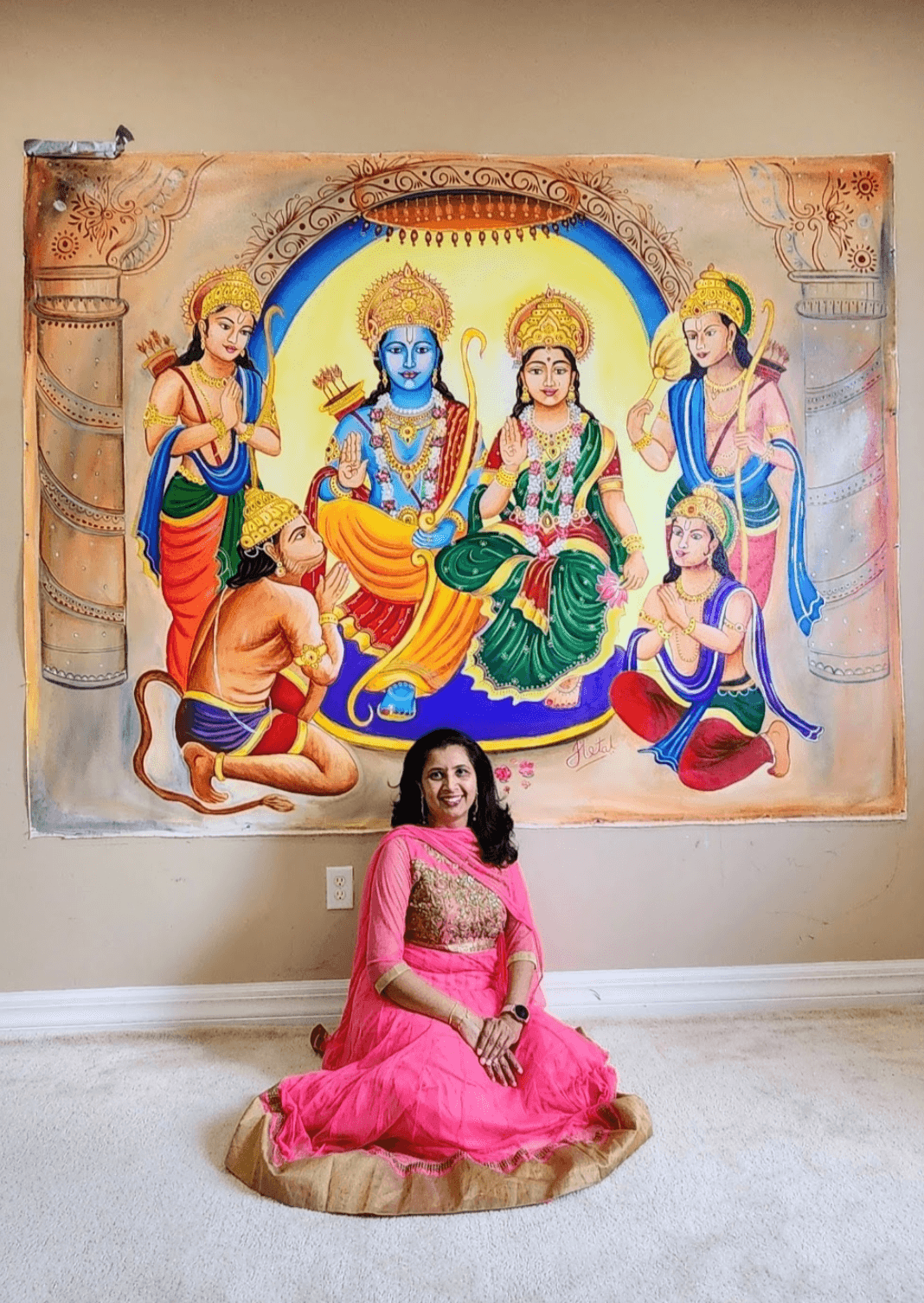 Woman in pink dress sitting on floor in front of colorful Hindu deity painting, with a beige wall background.
