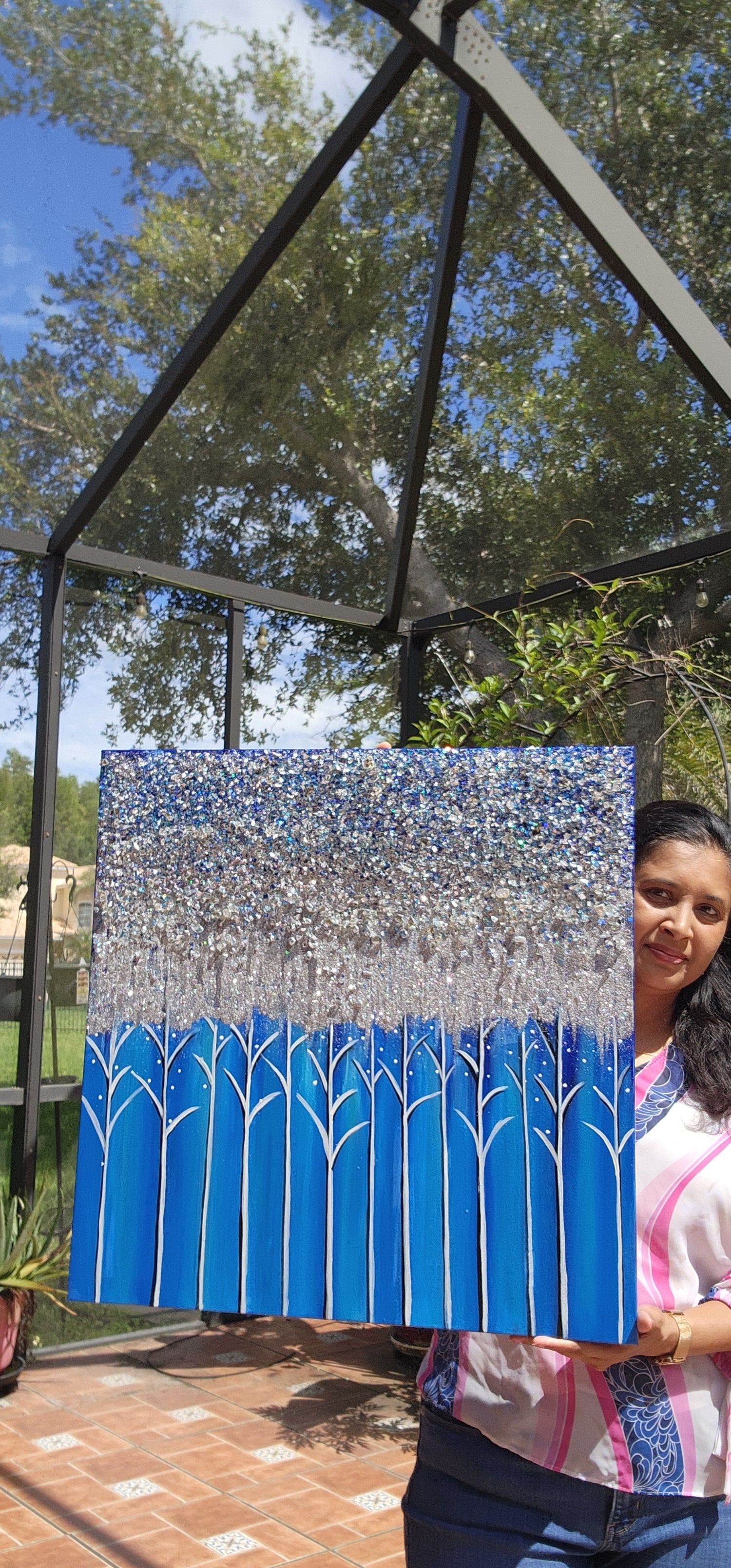 A person holding a glittery, silver rectangular object outdoors with trees and a blue sky in the background.