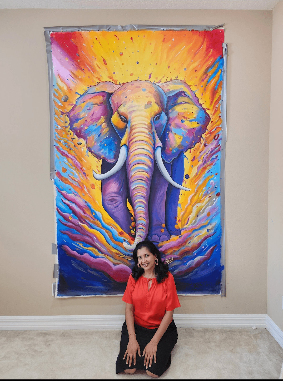 Woman sitting on floor in front of colorful elephant artwork on wall, smiling, wearing red top and black pants.