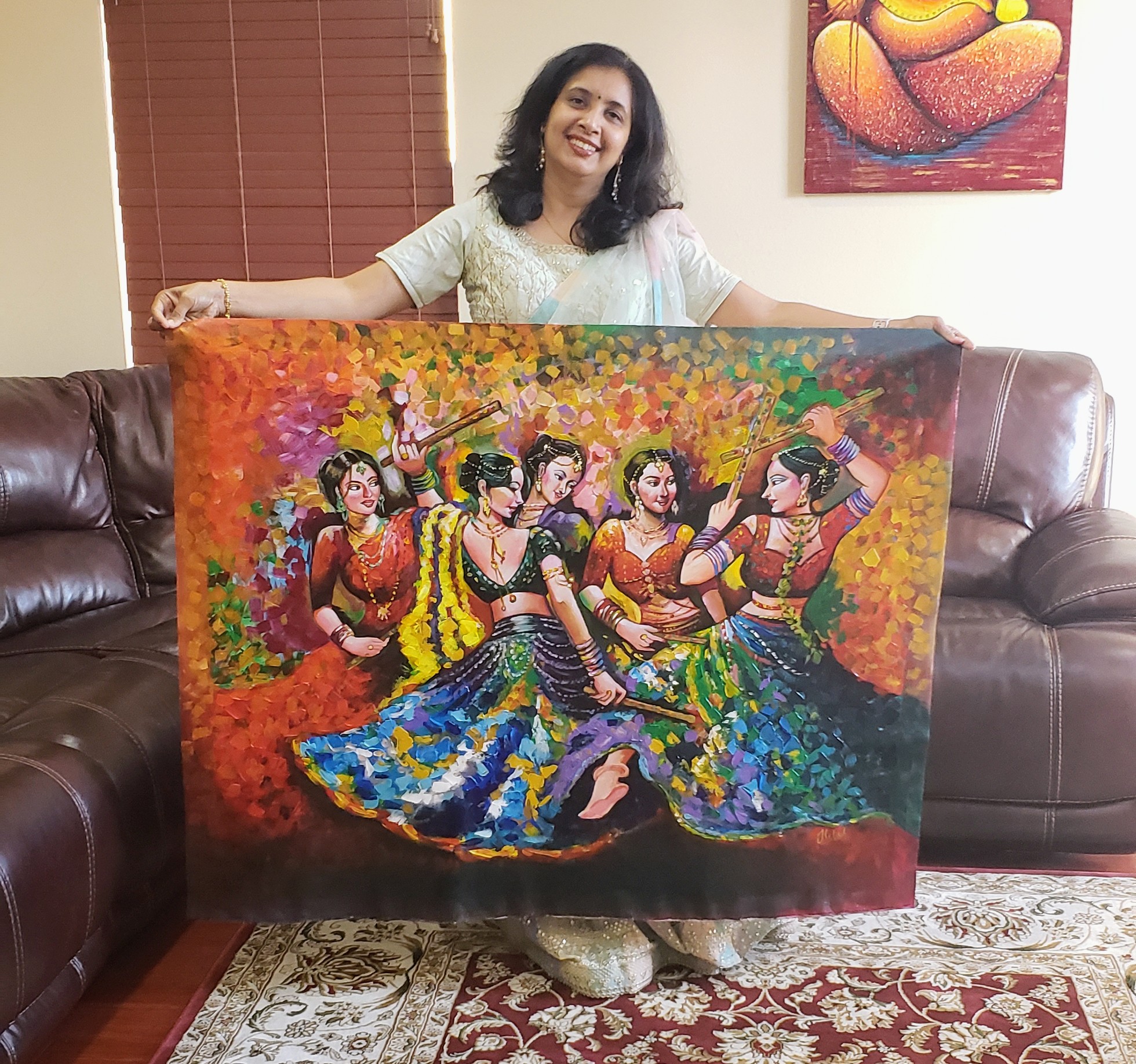 Woman holding a colorful painting of six women dancing outdoors, sitting on a sofa in a living room.