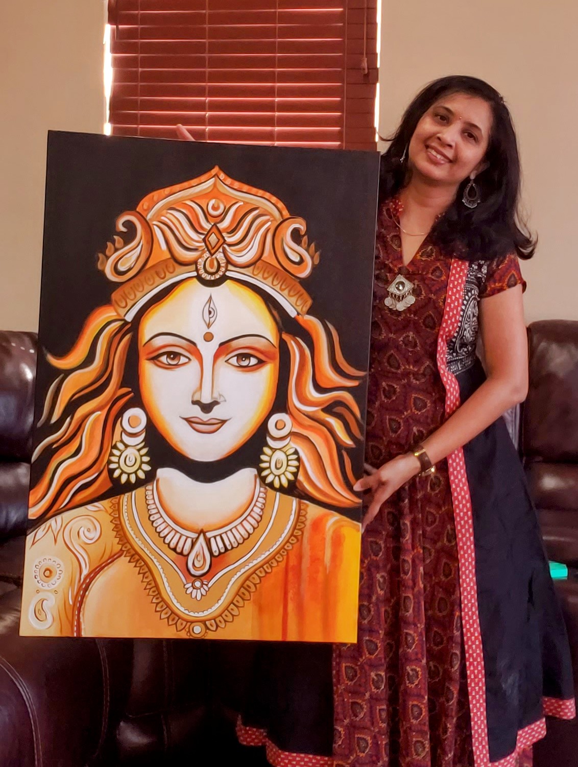 Woman standing next to a large painting of a goddess with orange and white features, wearing jewelry and a crown, indoors.