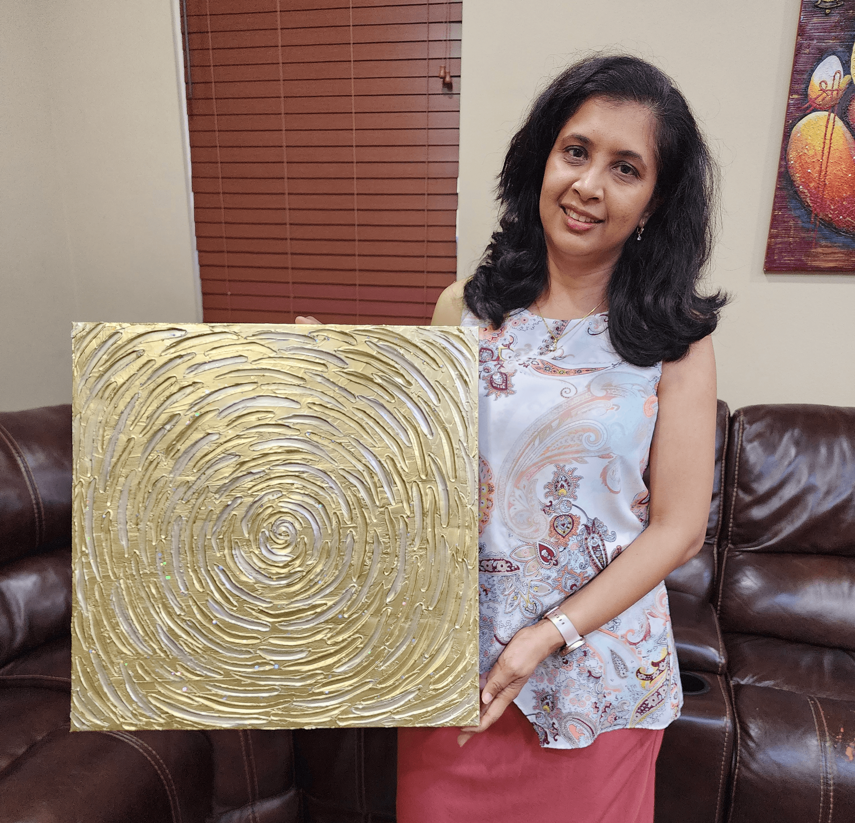 Woman with dark hair holding a gold textured square artwork, sitting on a brown leather couch, with a colorful painting on the wall behind.