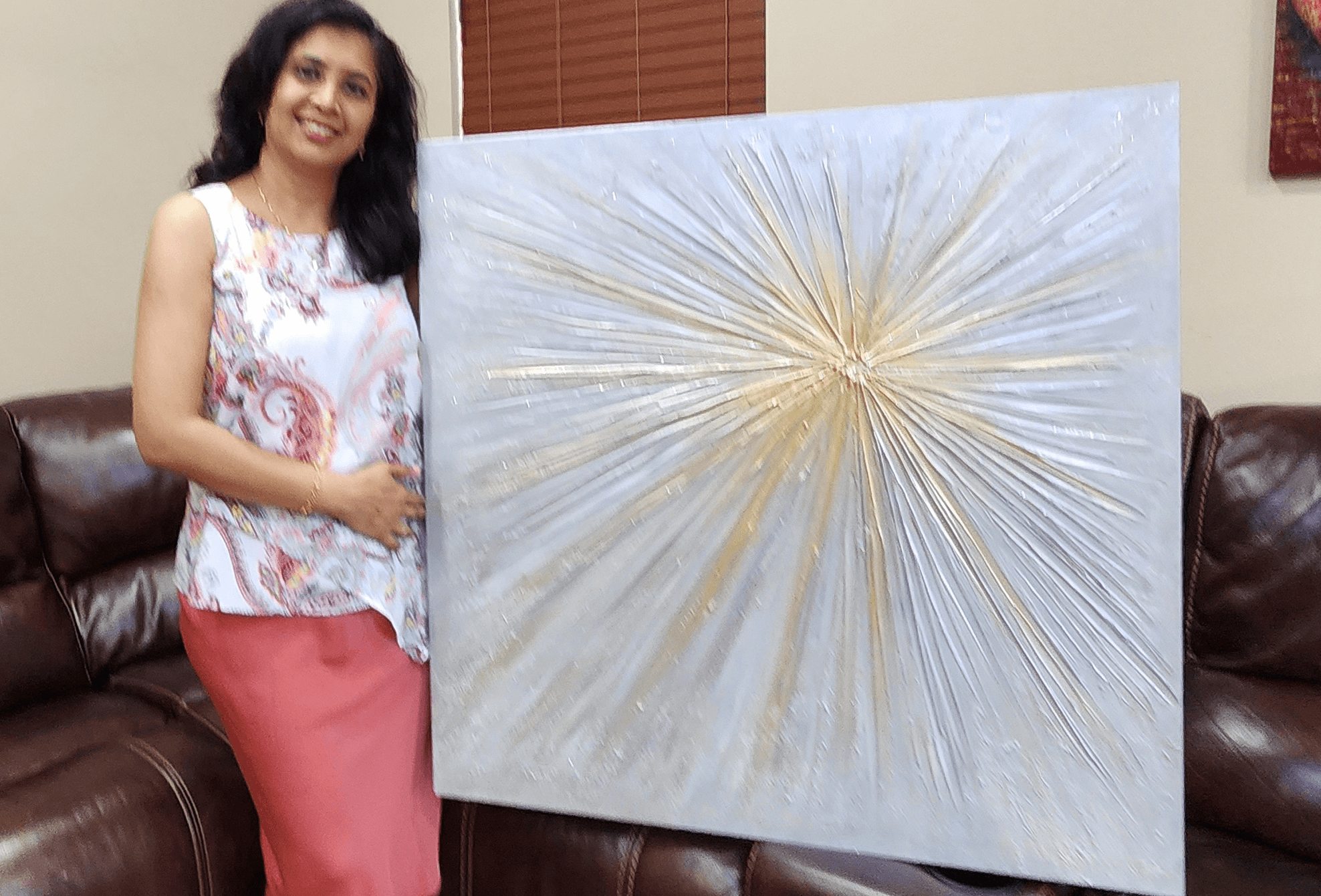 Woman standing next to large abstract painting with radiating gold lines on a white background, indoors.