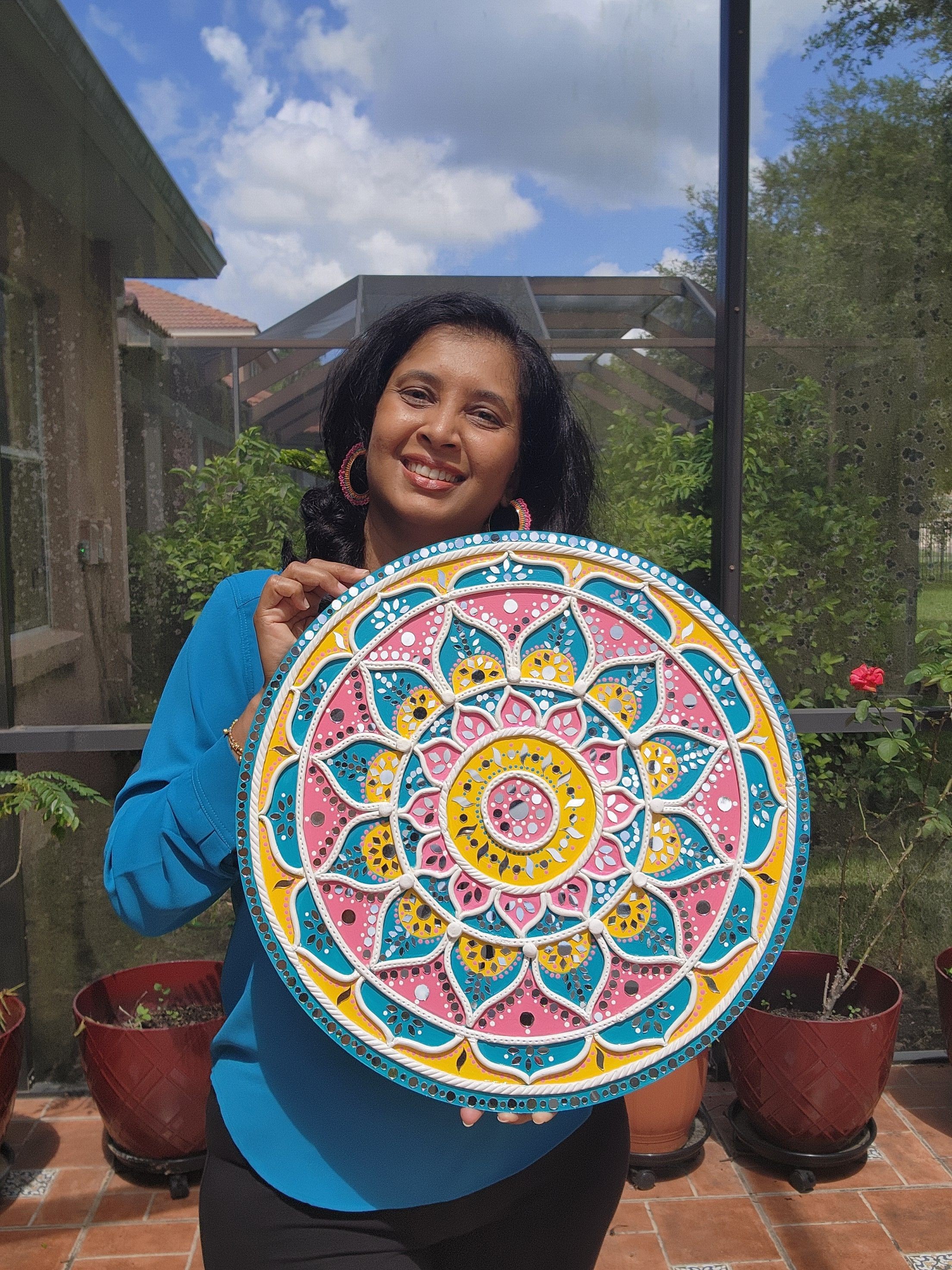Person holding a colorful circular mandala design, smiling outdoors with a blue sky and clouds in the background.