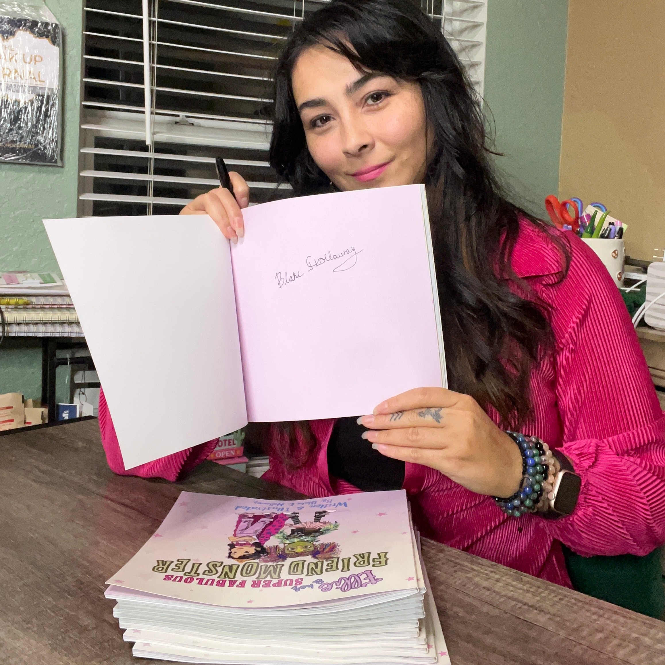 Woman with long dark hair holding open book with pink pages, smiling, sitting at desk with colorful supplies, books, and window behind.