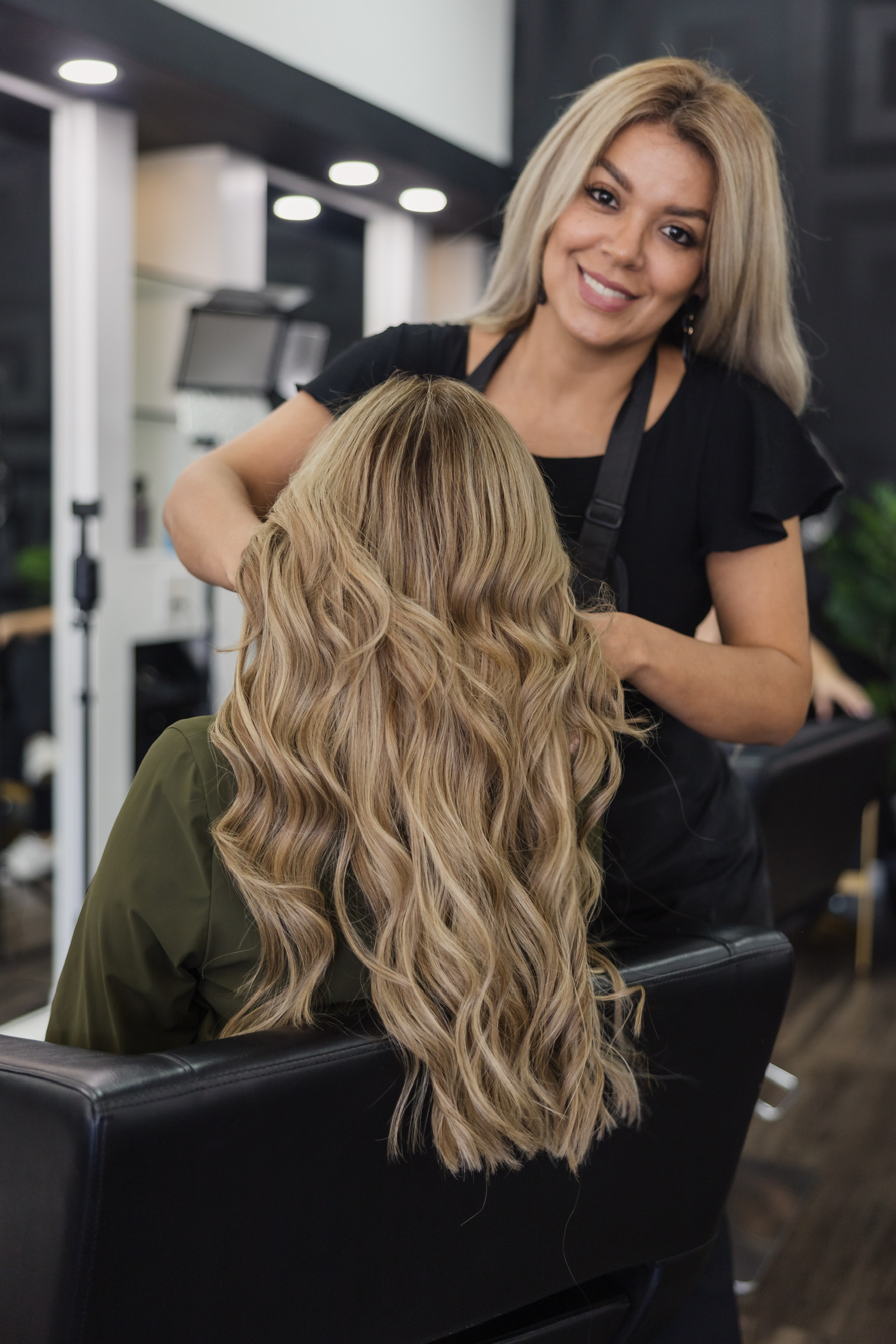 Hairdresser smiling at client with long, wavy blonde hair in salon setting.