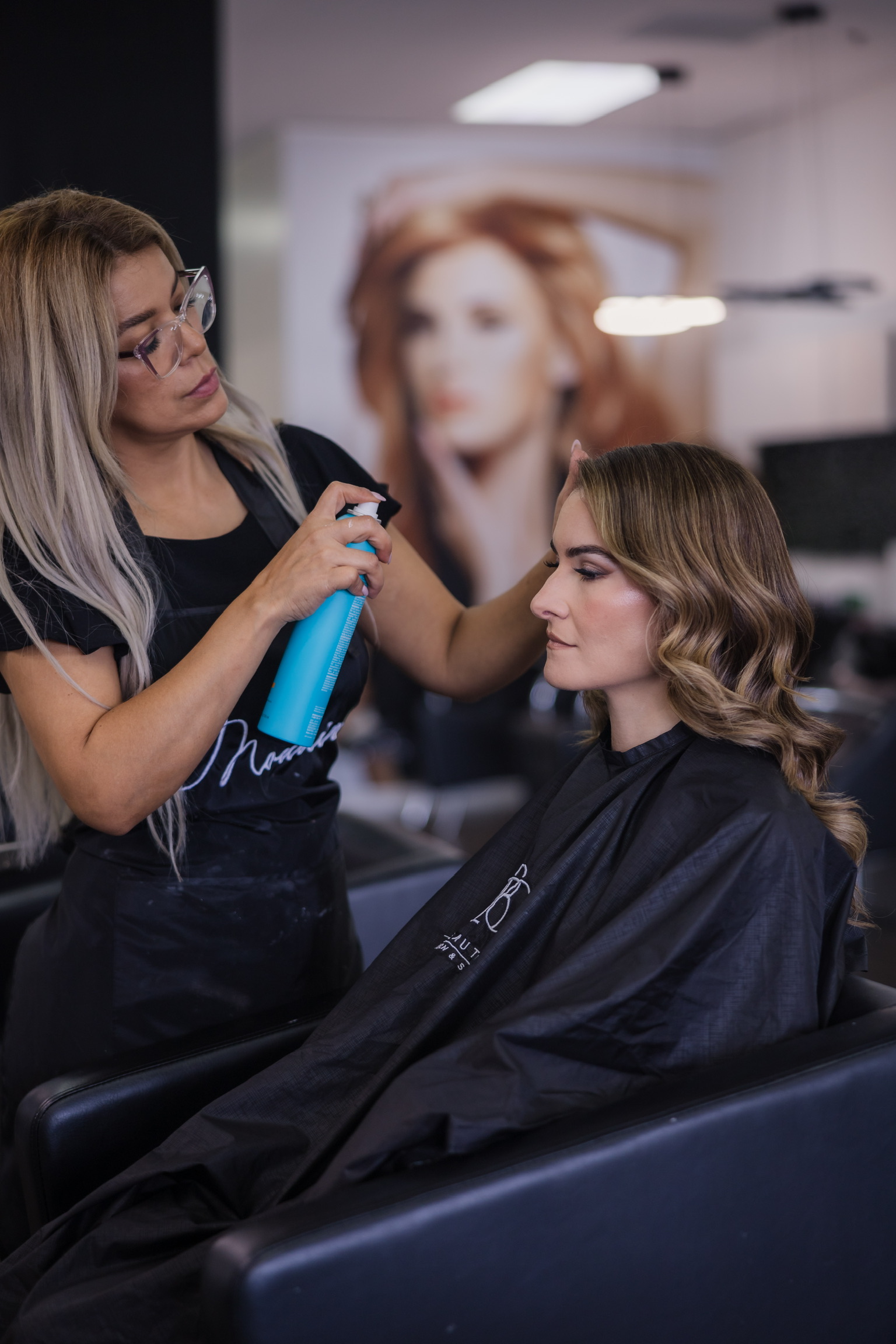 Hairdresser applies spray to a woman's hair in a salon, with a large portrait of a woman in the background.