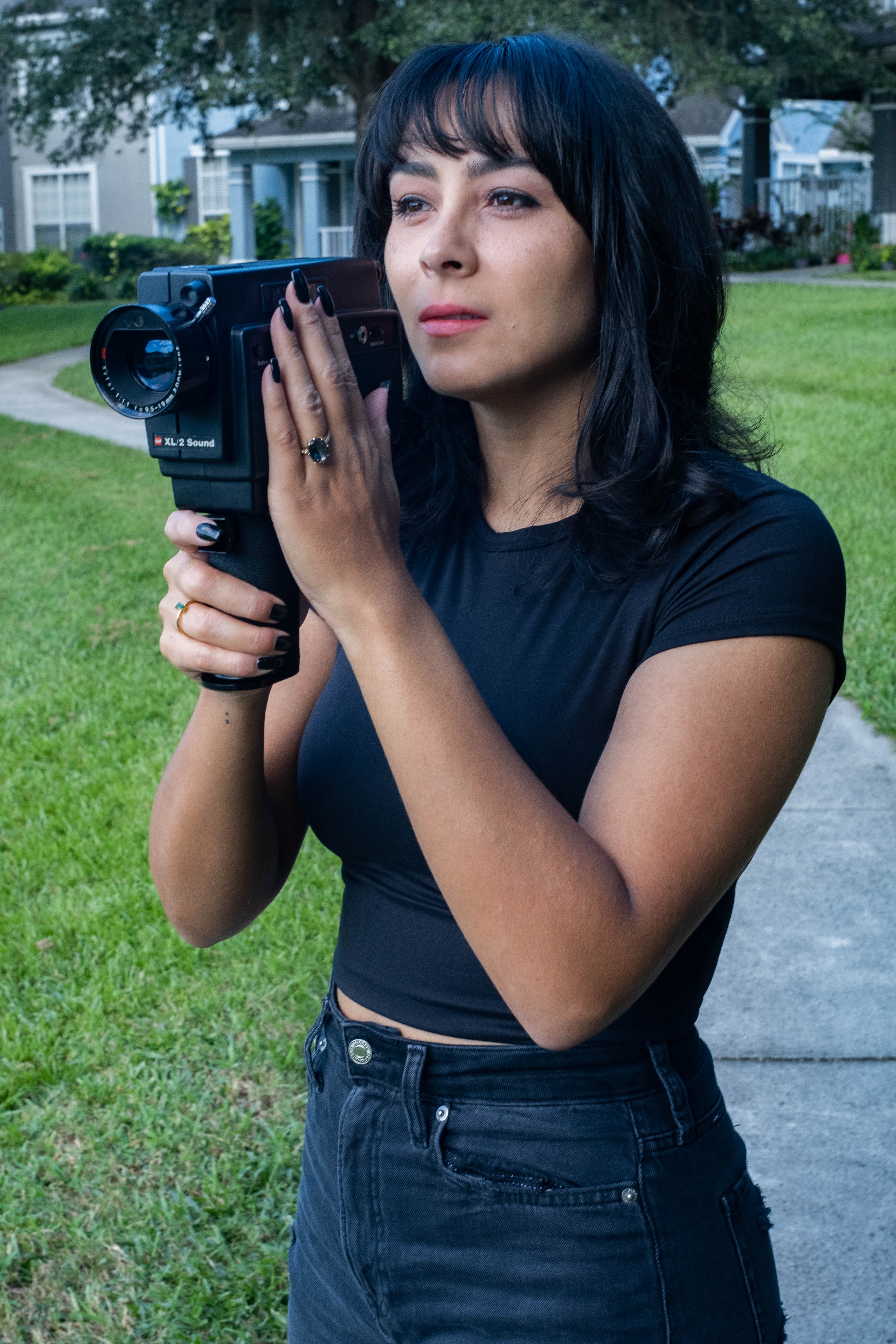 Woman with black hair holding a video camera outdoors on a grassy area, wearing a black shirt and jeans.