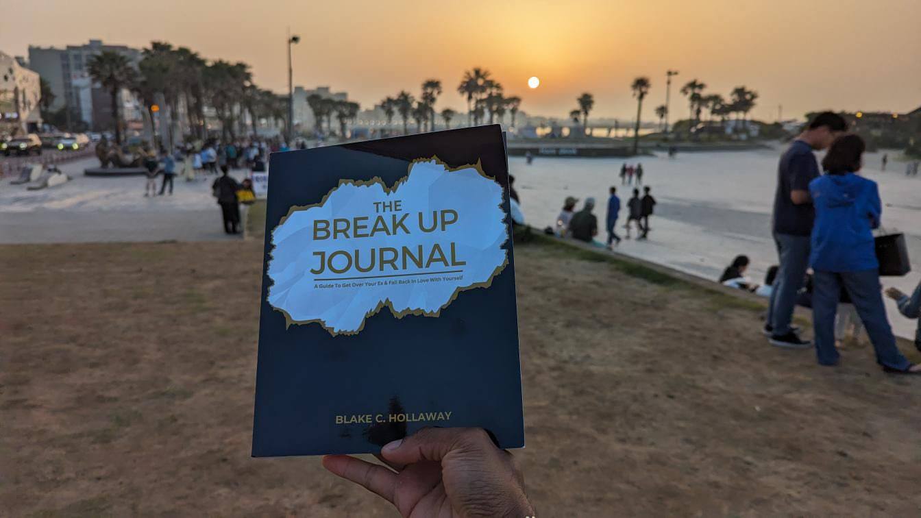 Hand holding a book titled 'The Breakup Journal' by Blake C. Holloway at a beach park during sunset.