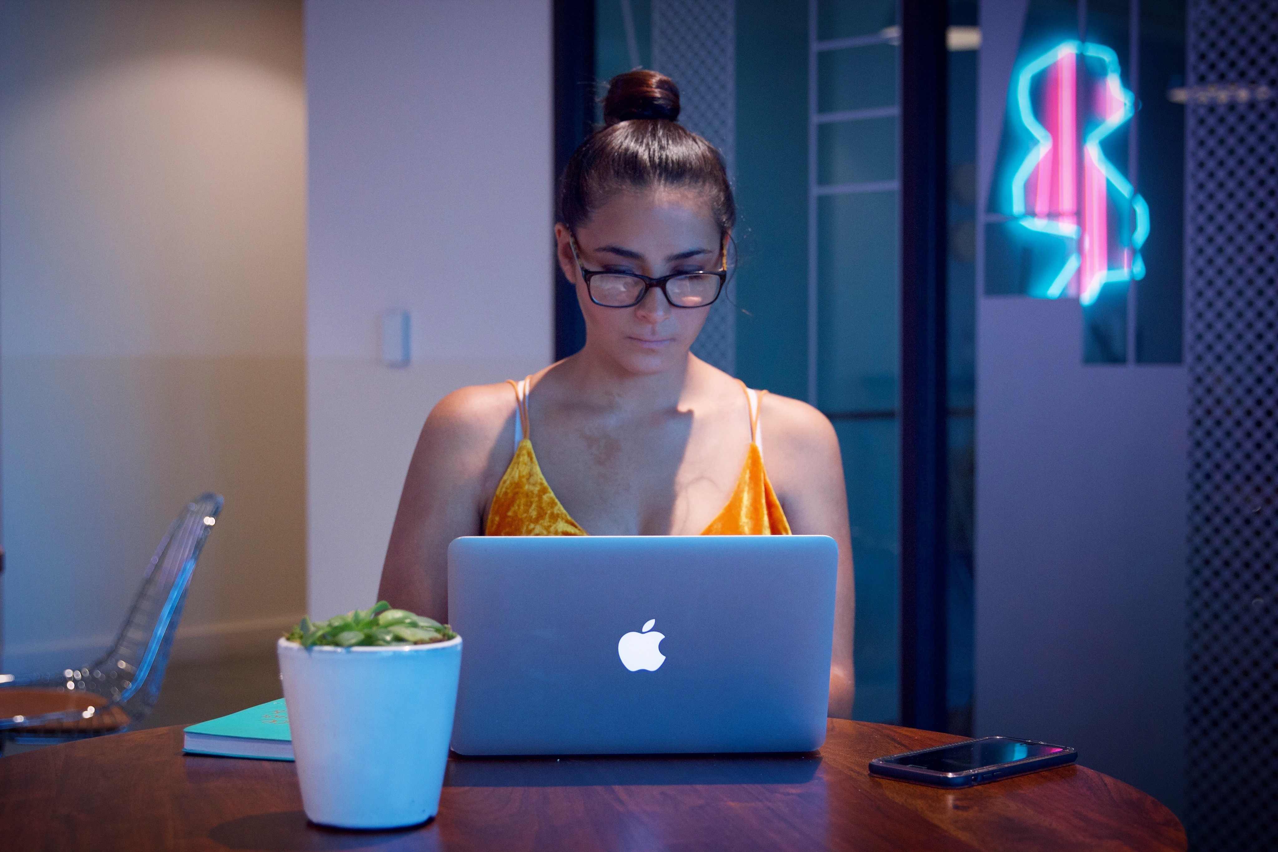 Woman with glasses working on a laptop at a table, neon sign in background, plant and phone nearby.