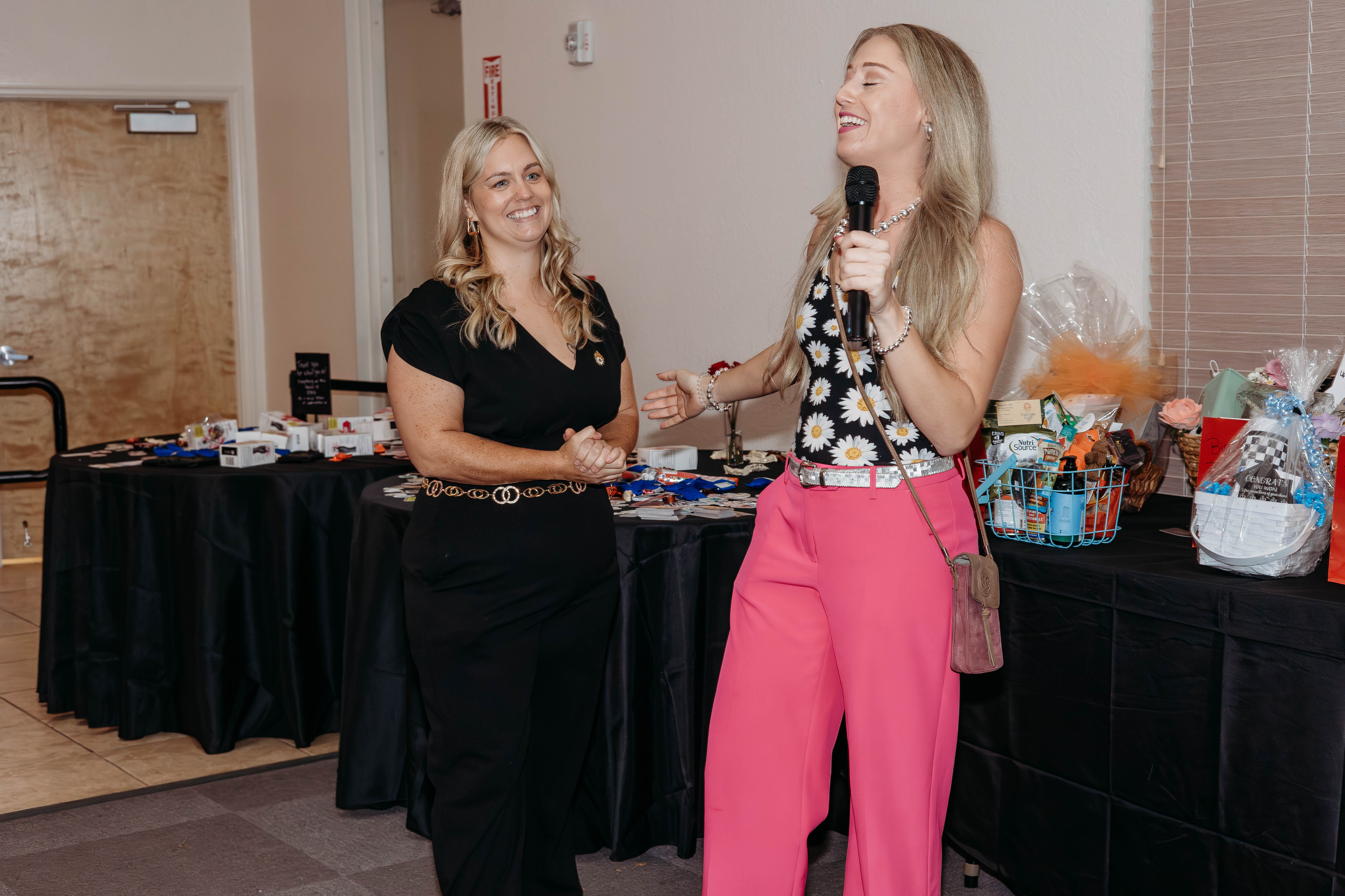 Two women standing and talking indoors, one holding a microphone, with a table of items behind them.