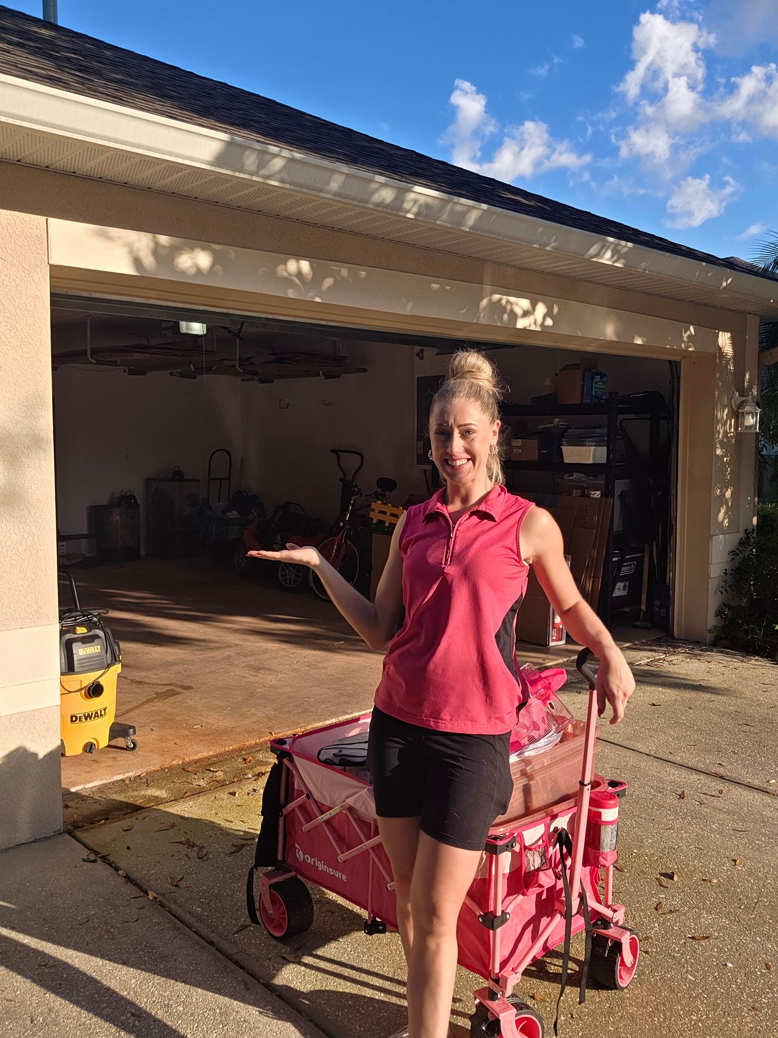 Woman in pink shirt and black shorts standing outside a building, with a pink stroller nearby, under a blue sky.