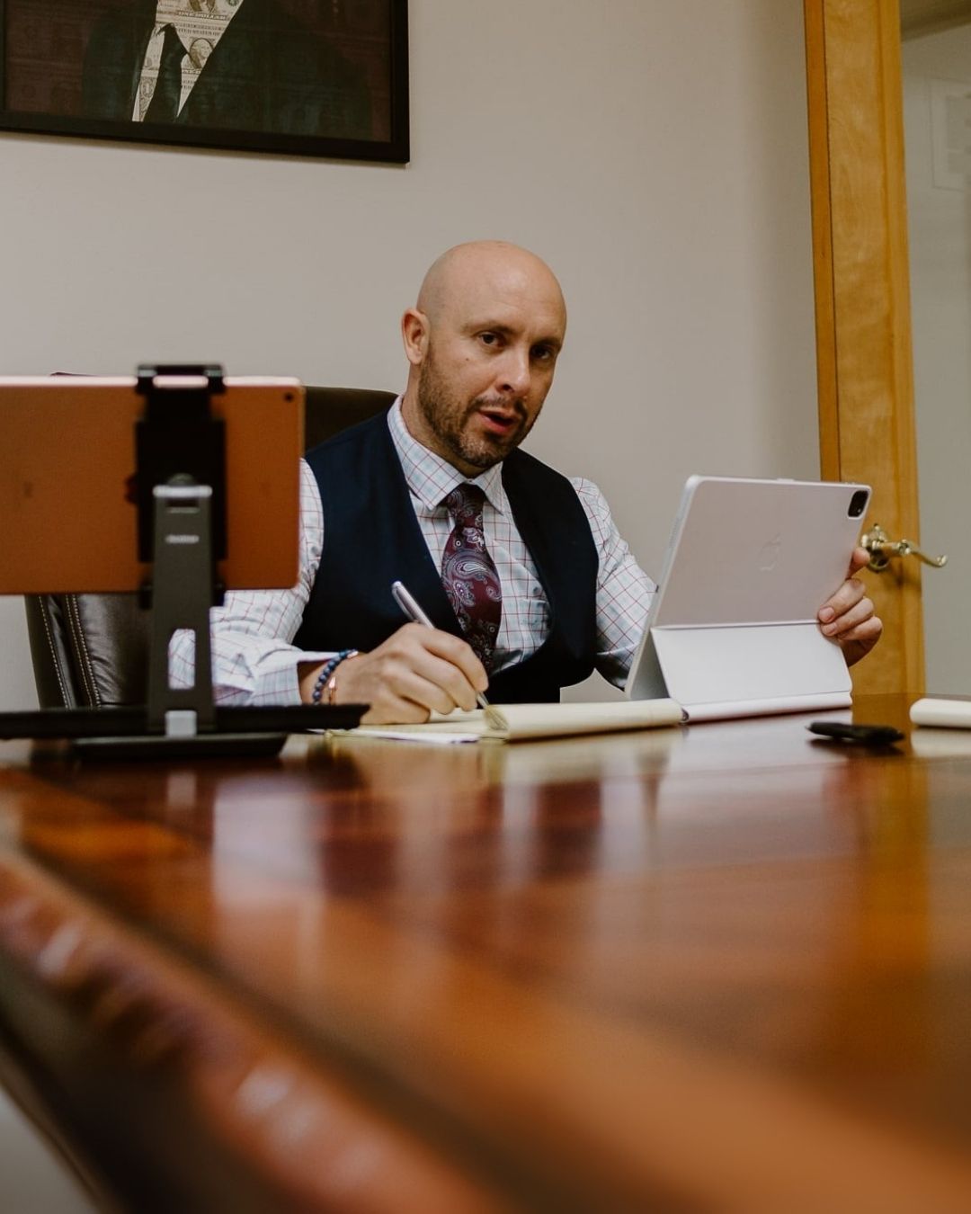 Bald man in a white shirt and tie, sitting at a desk with a tablet, in an office setting.