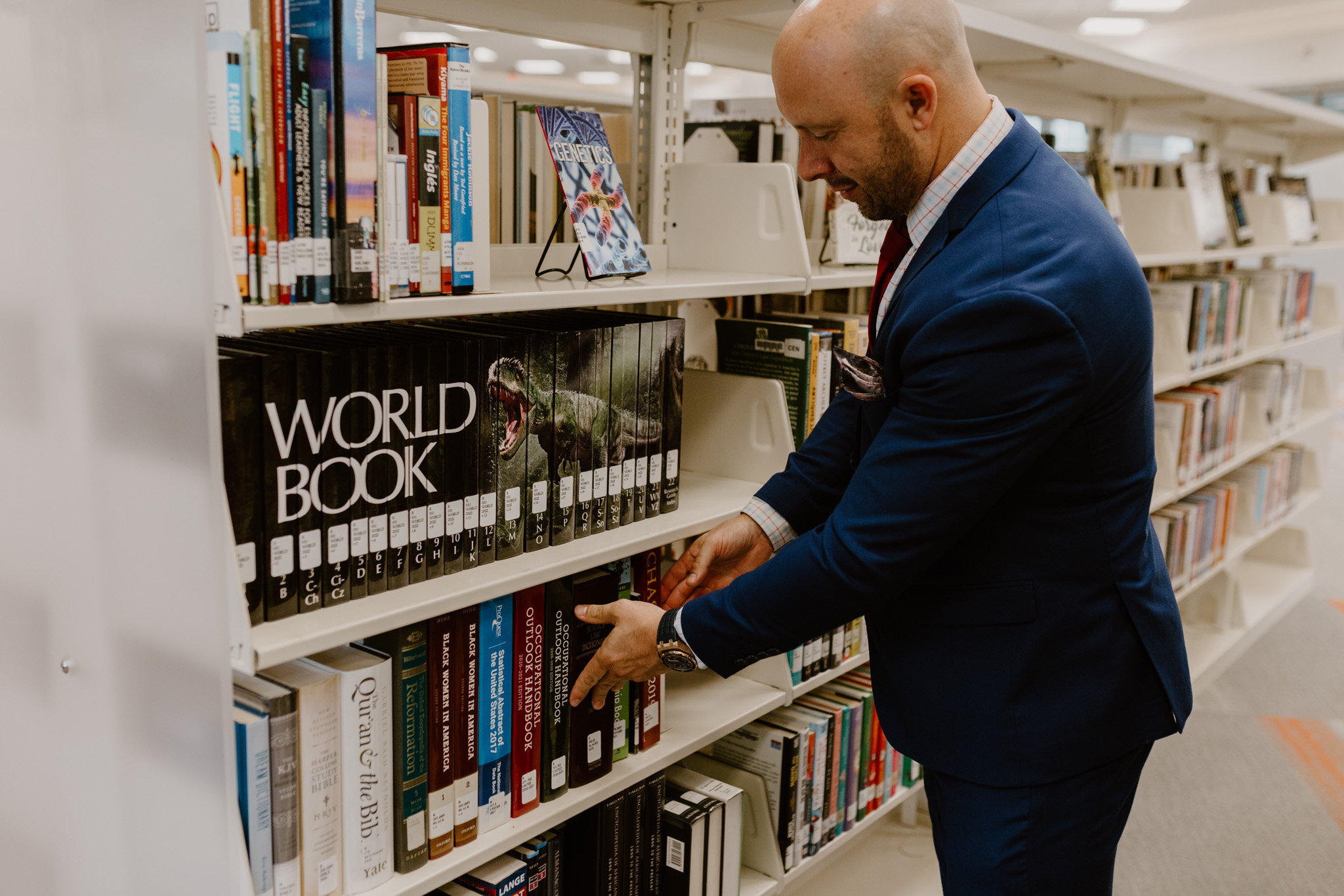 Man in a suit browsing books on a library shelf, with a 'World Book' sign visible, in a well-lit library.
