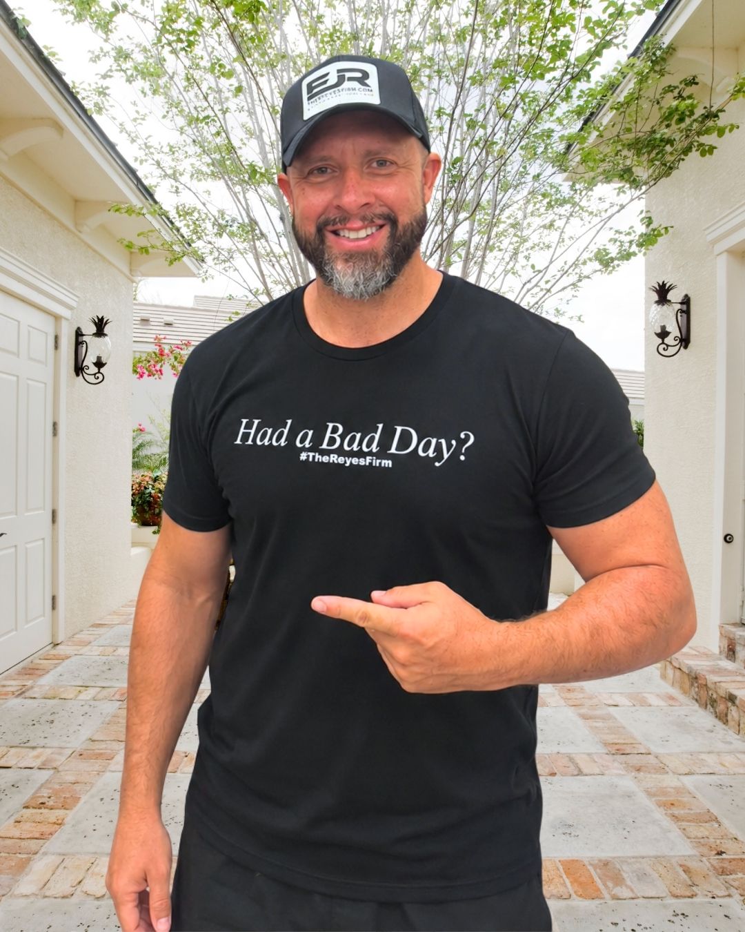 Man with a beard smiling, wearing a black T-shirt and cap, pointing at his shirt, standing outdoors with trees and buildings behind him.