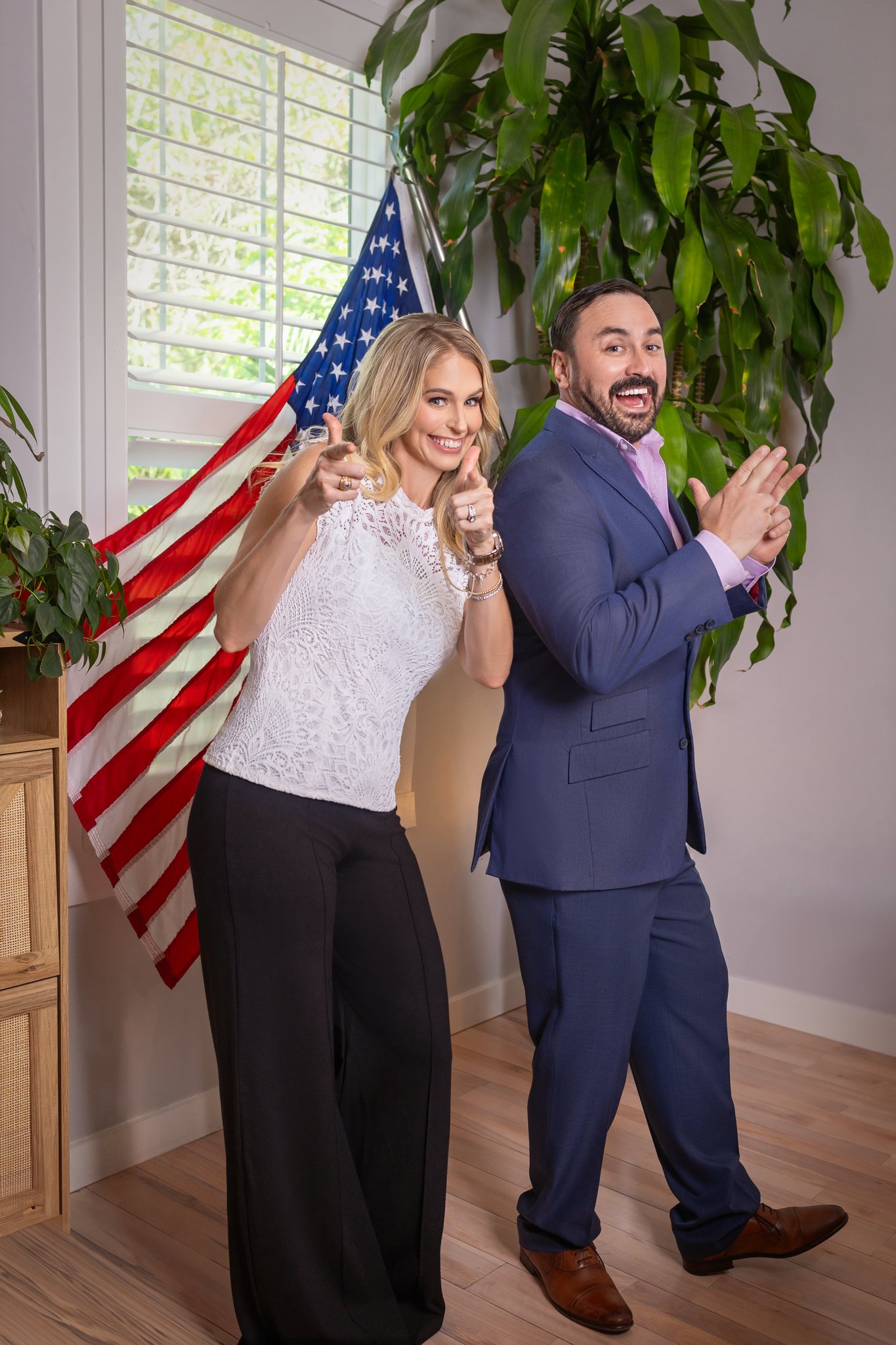 Two people celebrating indoors with an American flag, smiling and clapping, near a large plant and window.