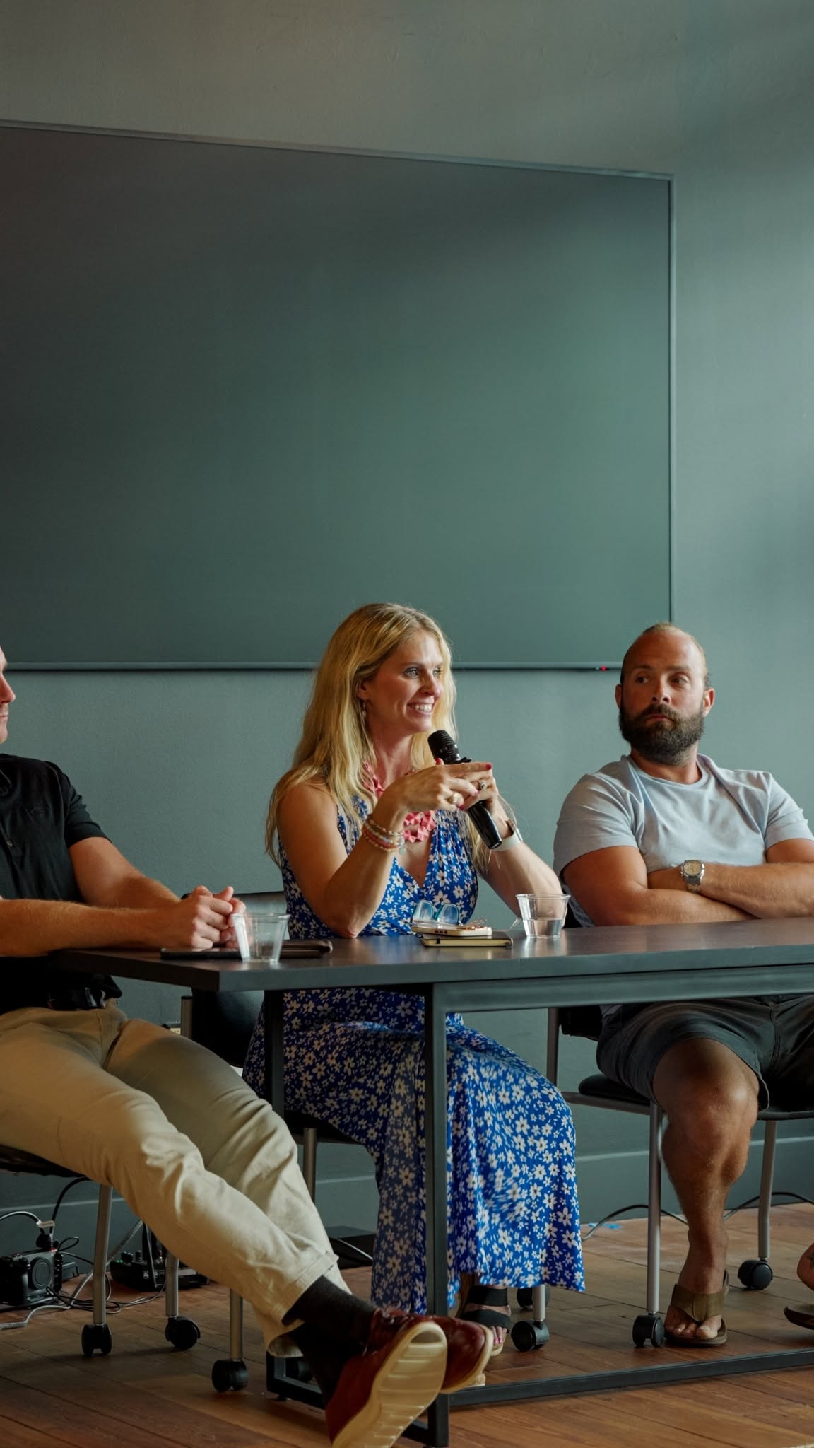 Three people sitting at a table, woman speaking into a microphone, two men listening, in a room with a large blank wall.