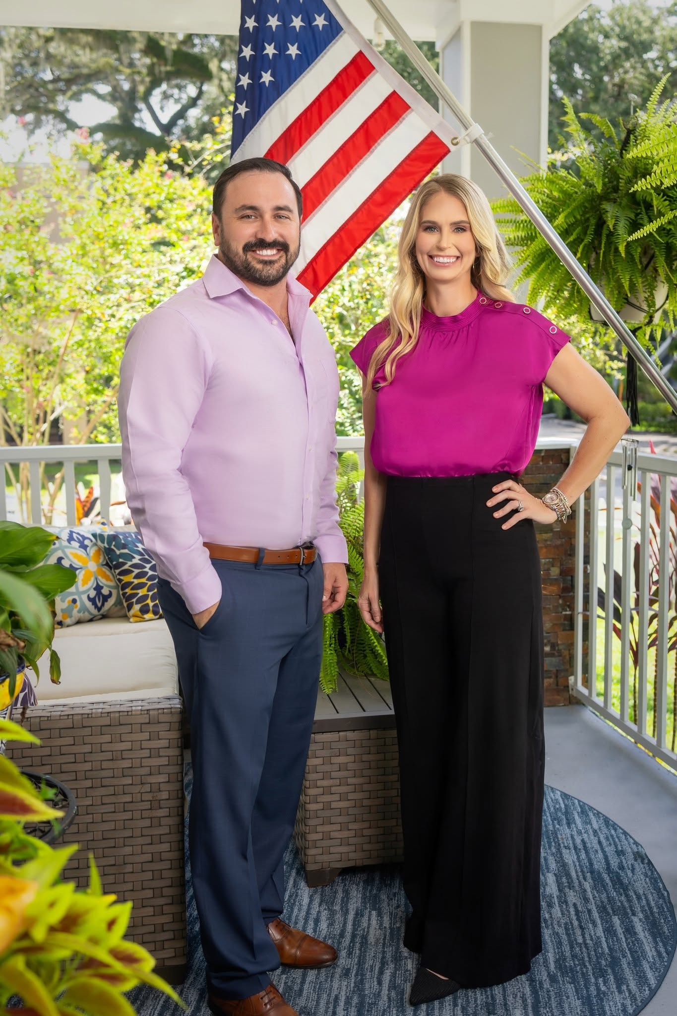 A man and woman stand on a porch with an American flag behind them, smiling at the camera.