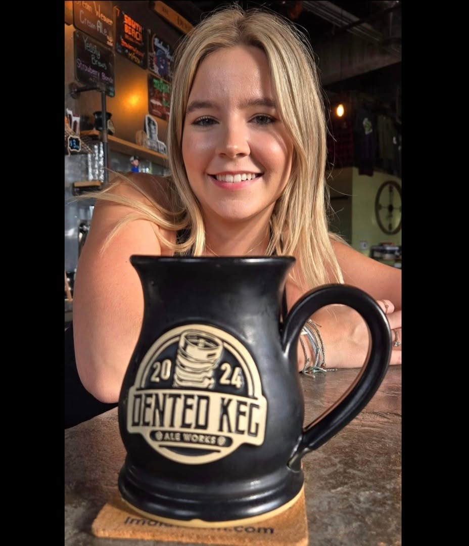Young woman with blonde hair smiling at a bar, holding a black mug with a logo, in a dimly lit setting.