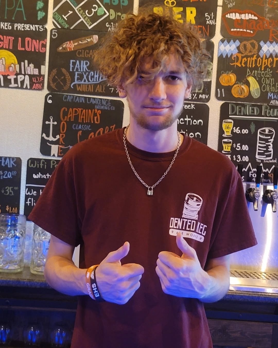 Young man with curly hair giving thumbs-up, standing in front of a colorful chalkboard menu, wearing a maroon T-shirt and necklace.