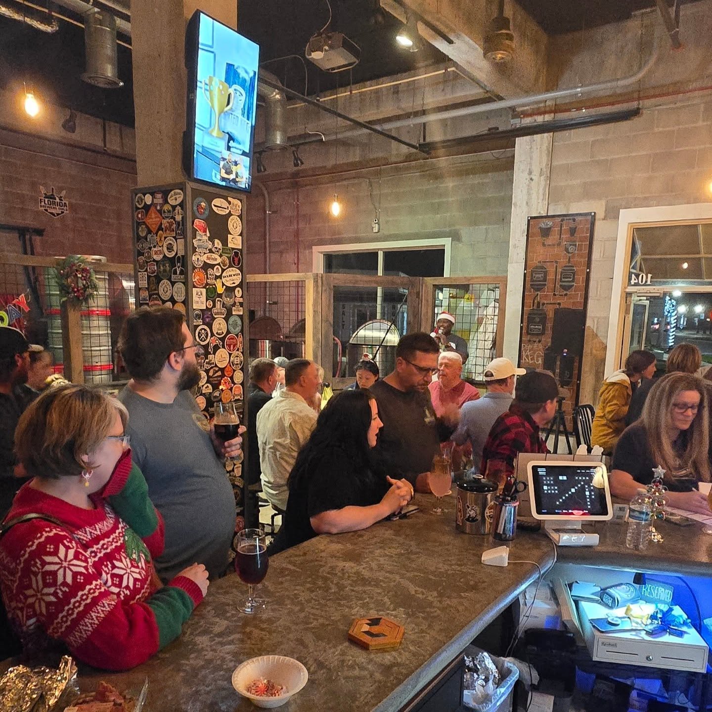 People gathered around a bar counter in a lively indoor setting with brick walls and industrial decor.