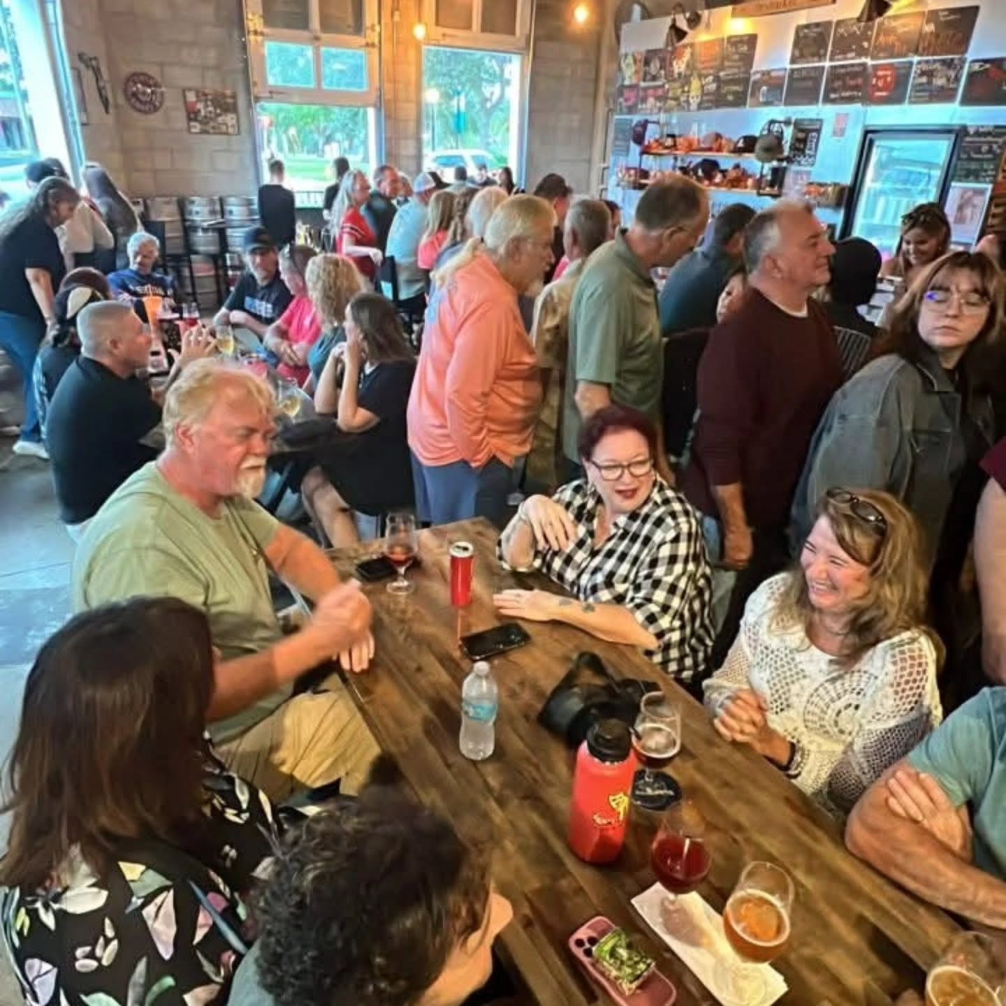 Crowded indoor space with people sitting and standing, some at a long table, others near windows and walls, lively atmosphere.