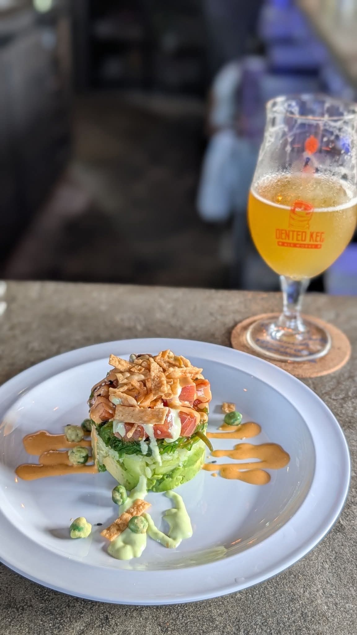A plated dish with a layered salad topped with a creamy sauce, garnished with avocado slices, and a glass of beer in the background.