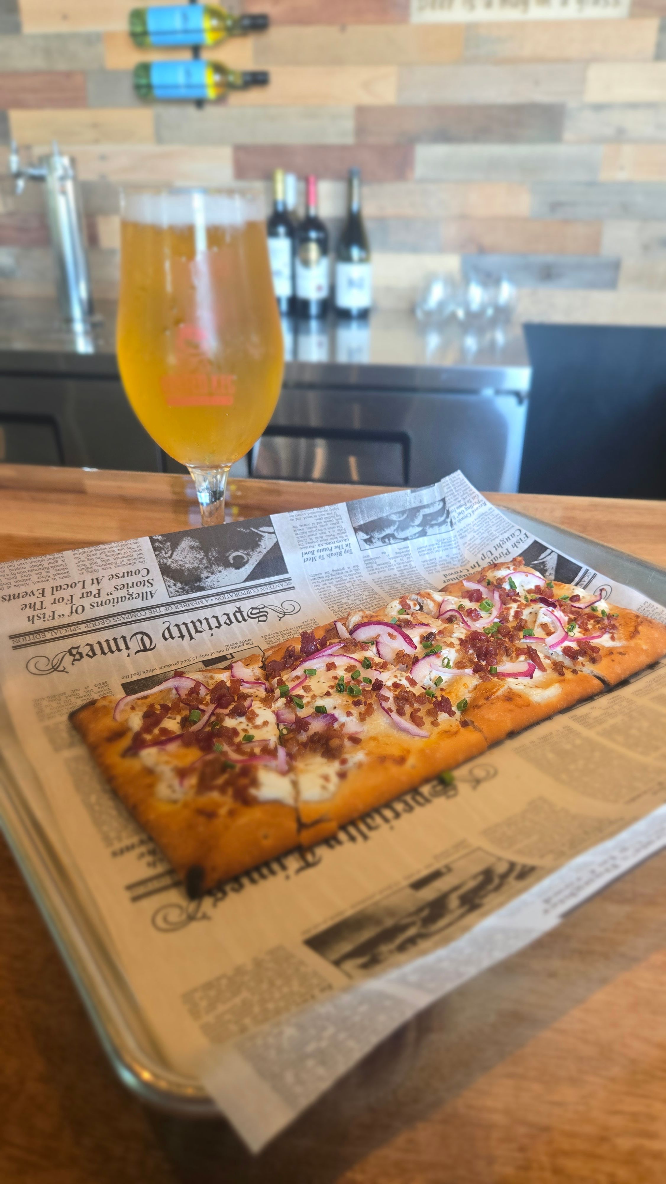 A slice of pizza on newspaper, a glass of orange juice, and bottles on a wooden table.
