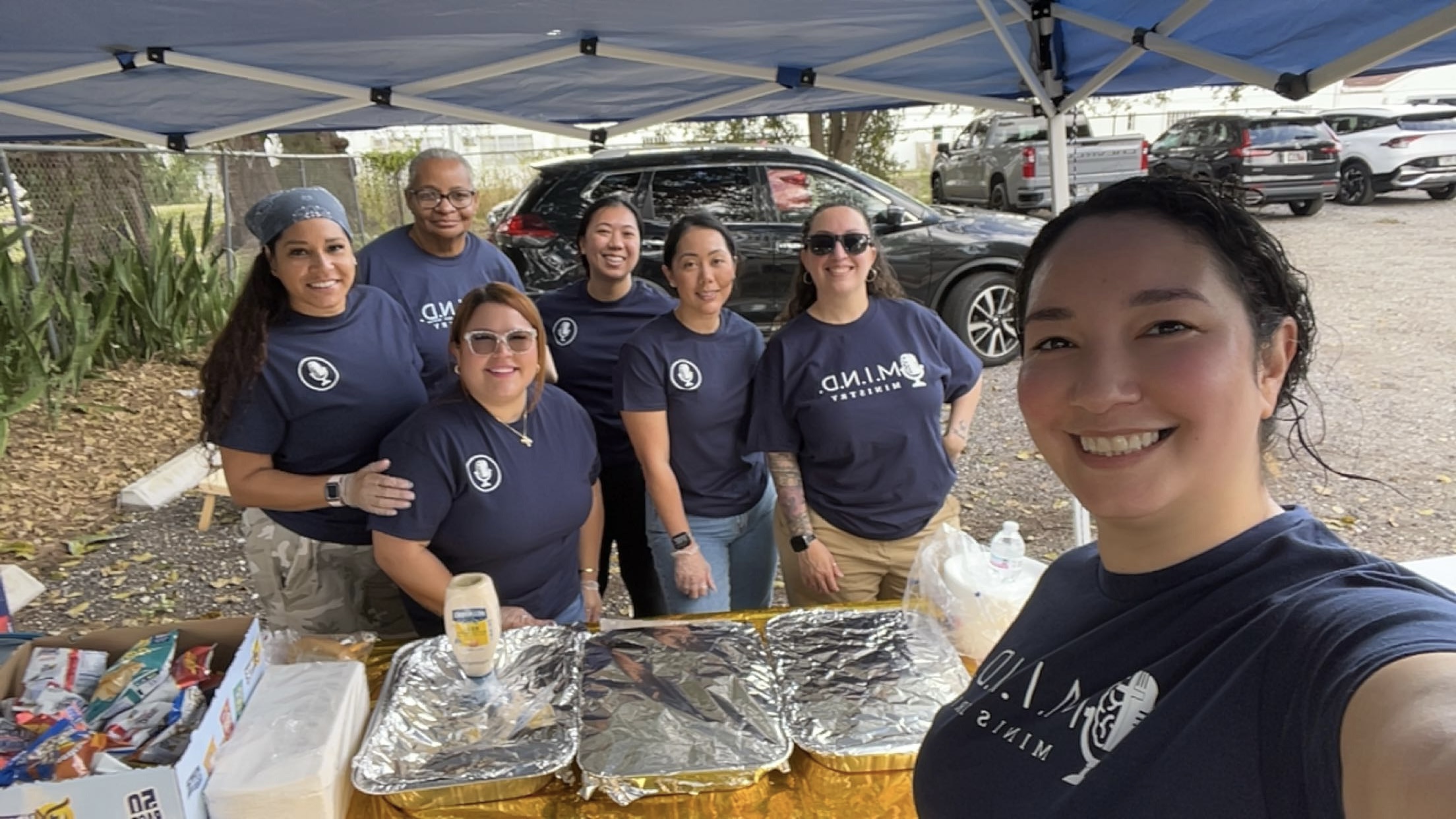 Group of seven people wearing matching shirts under a blue canopy, smiling at the camera, with food trays in front.